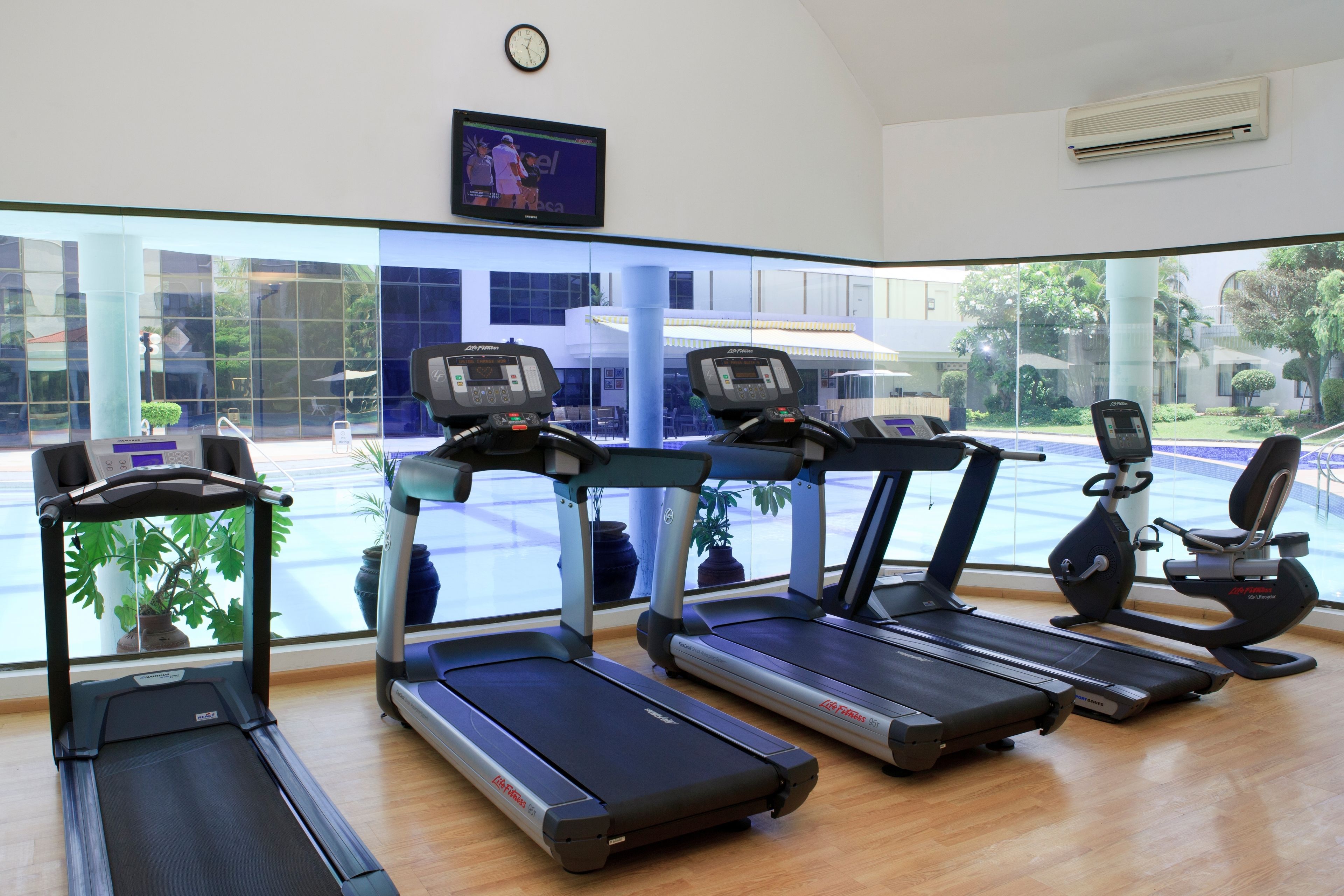 A gym with treadmills, a stationary bike, and a rowing machine. There are large windows with a view of buildings and trees.