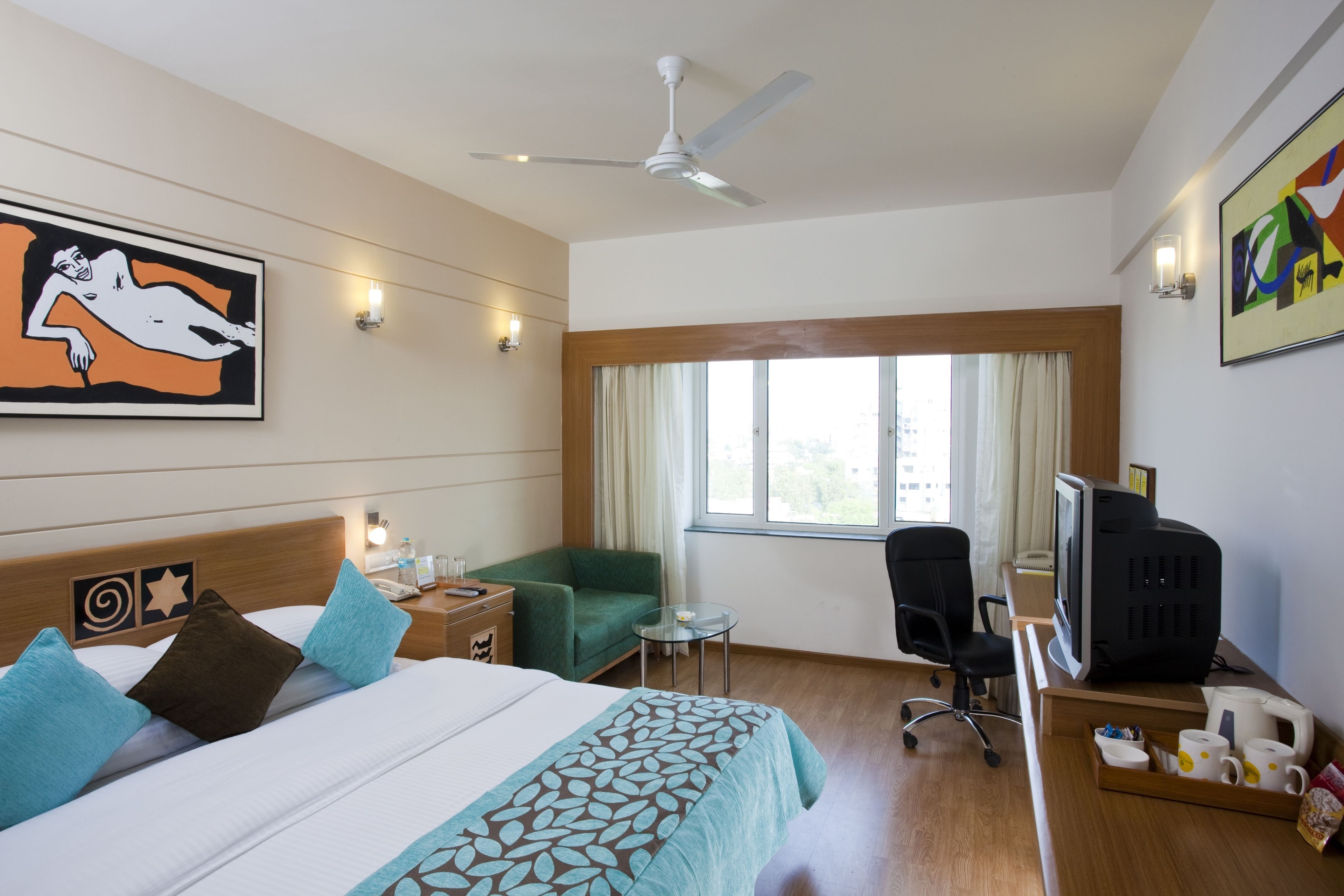 Habitación de hotel con cama, escritorio, computadora, silla, mesita y ventana con cortinas.
