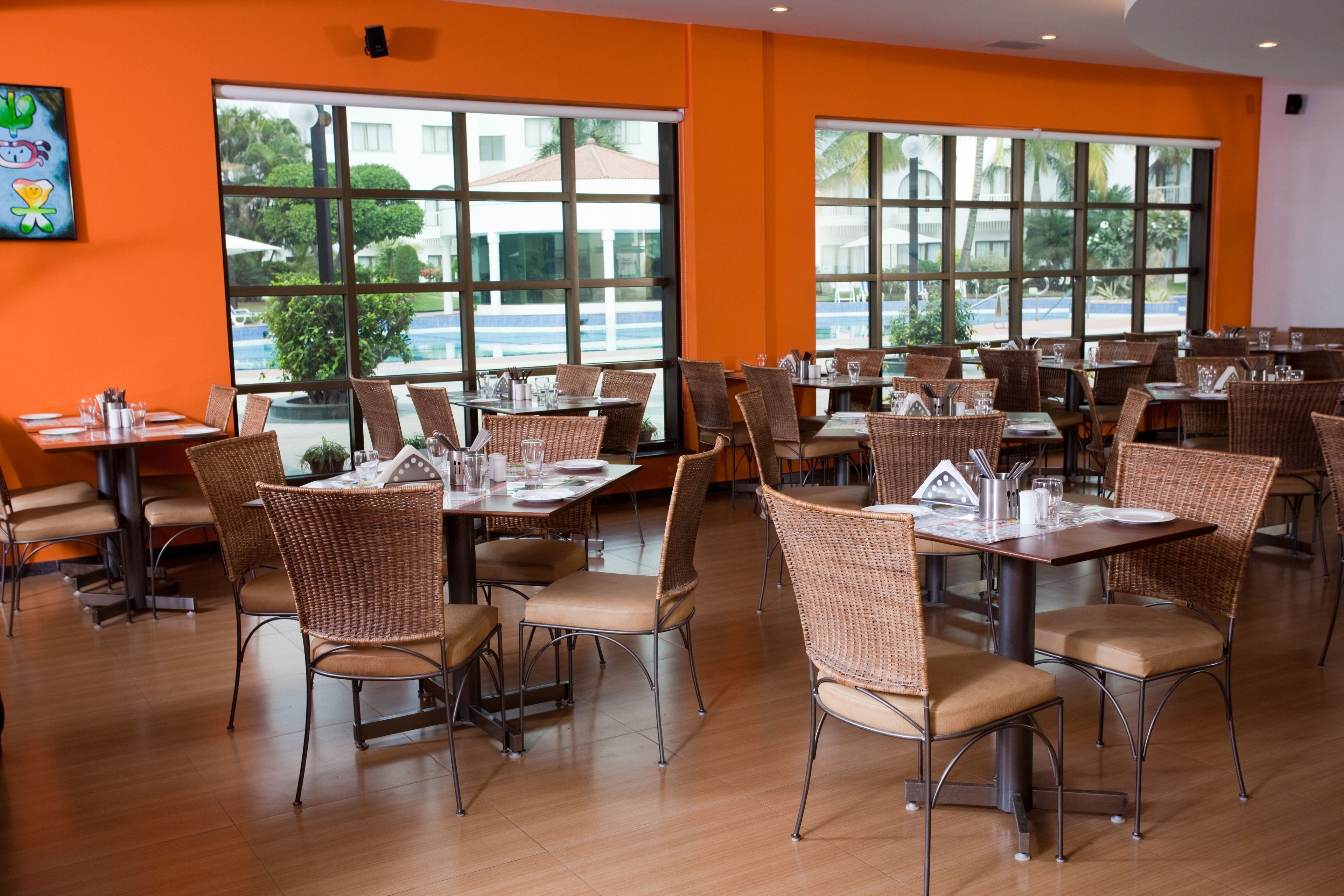A restaurant with multiple tables and wicker chairs, large windows with a view of palm trees and buildings, and orange walls.