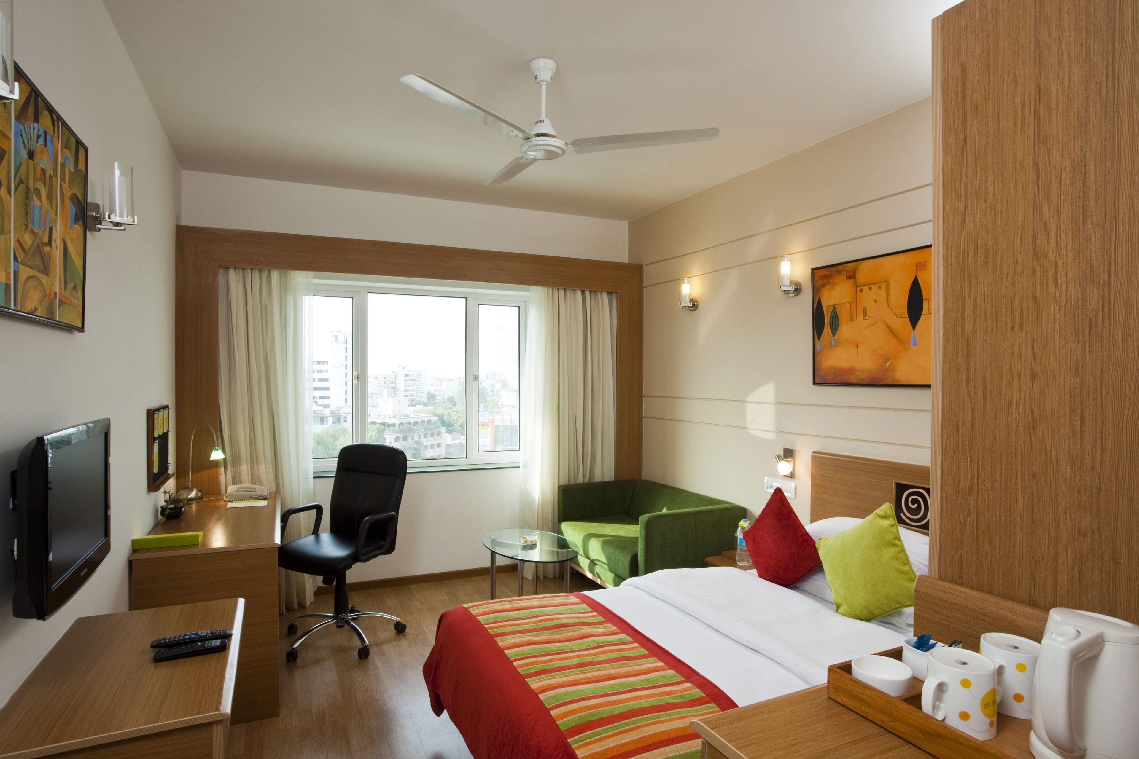 A hotel room with a bed, a desk, a chair, a television, a ceiling fan, a window with curtains, and artwork on the walls.