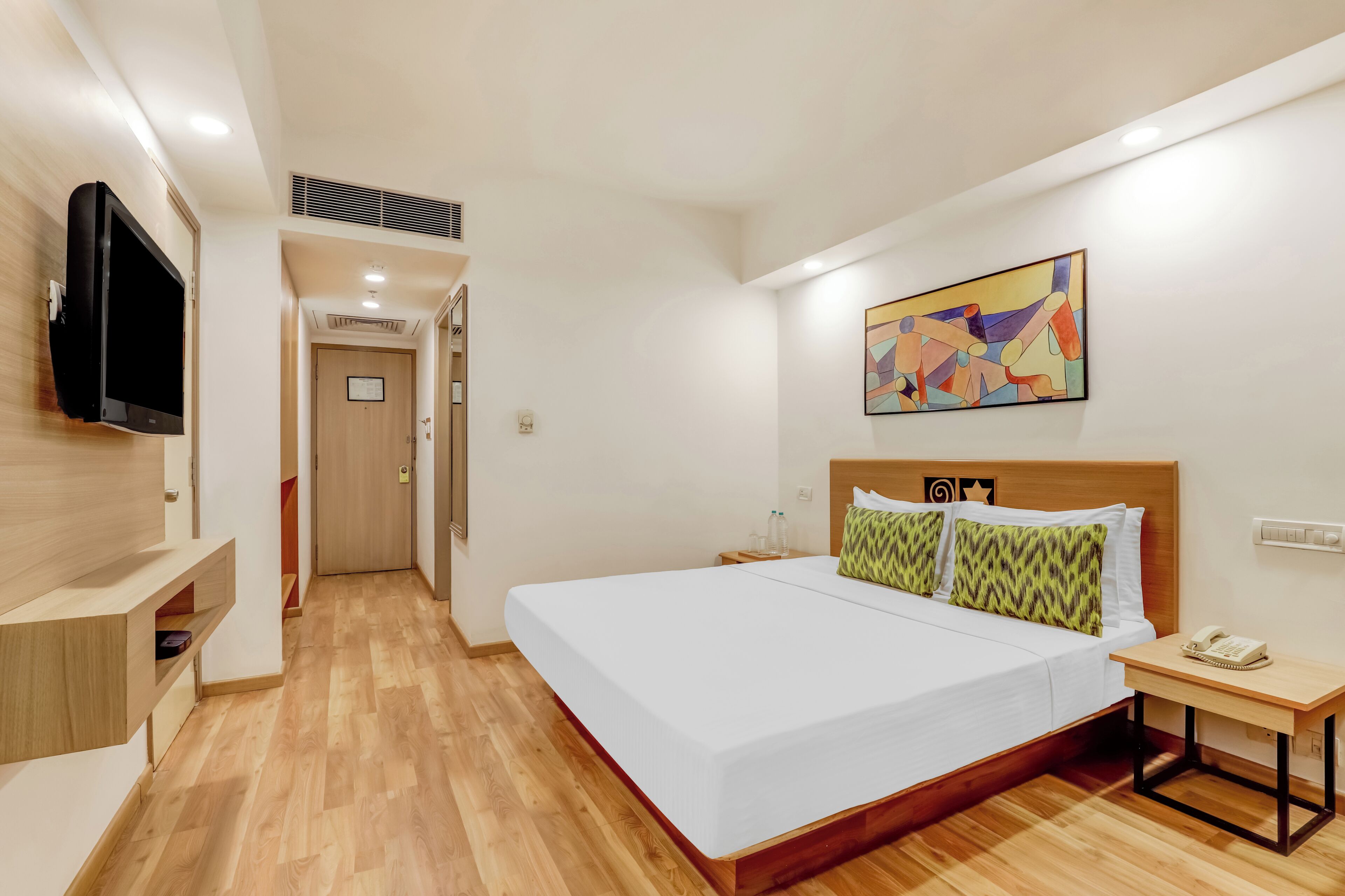 A hotel room with a large bed, a bedside table with a phone, a TV mounted on the wall, and a colorful abstract painting above the bed.