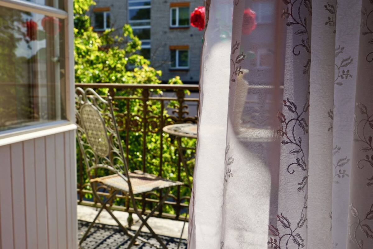 Parvekkeella on tuoli, pöytä ja ikkuna, josta avautuu näkymä vehreään maisemaan ja rakennuksiin.