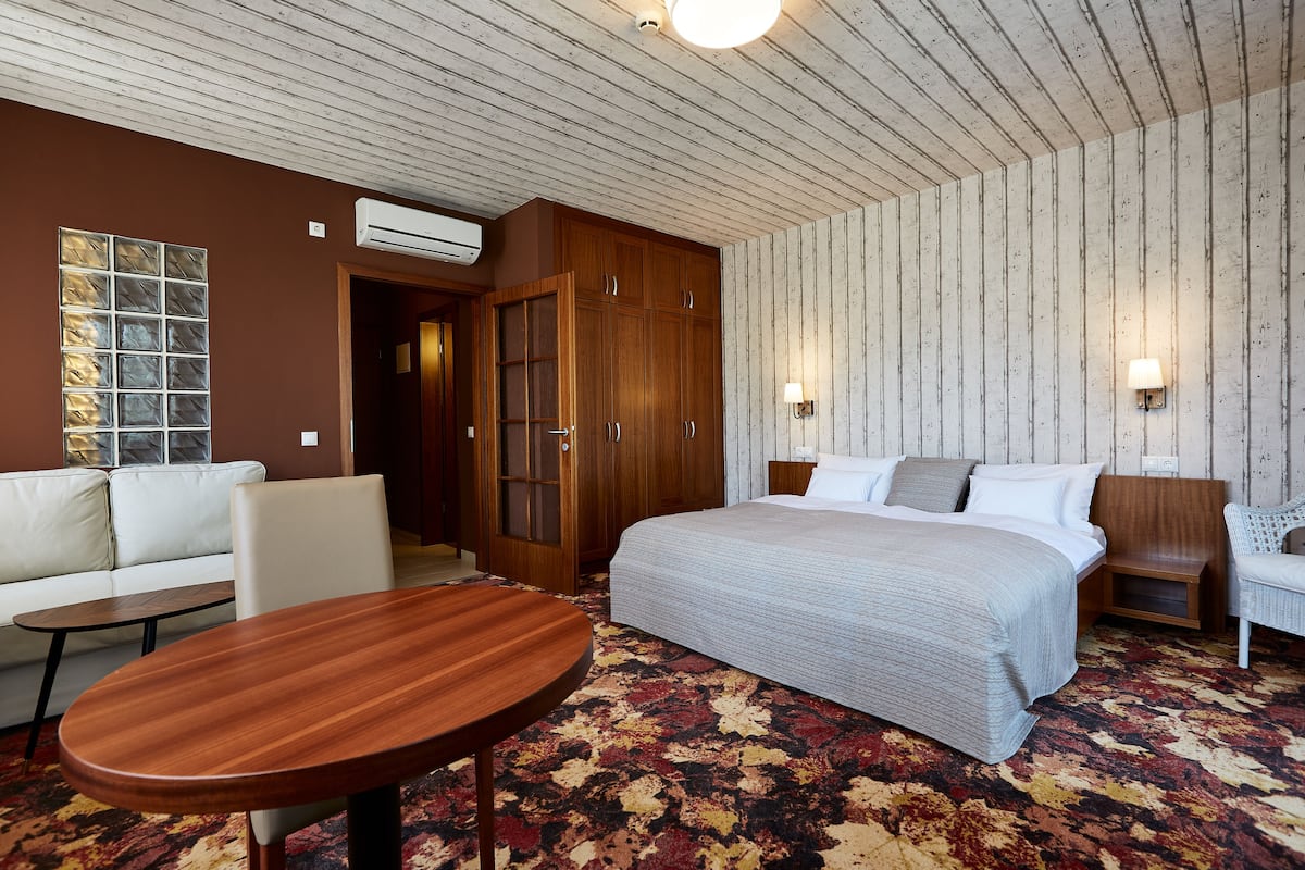 A hotel room with a large bed, a wooden wardrobe, a sofa, a small table, and a patterned carpet.