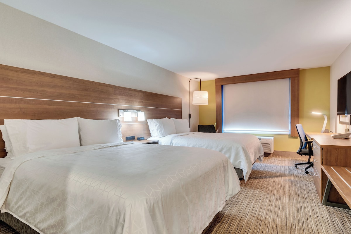 Quarto de hotel com duas camas, uma escrivaninha, uma cadeira e uma janela grande.
