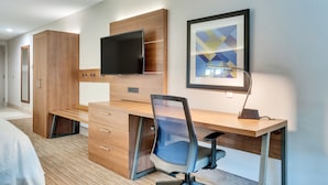 A modern hotel room with a wooden desk, a flat-screen TV, a framed abstract artwork, and a bed.