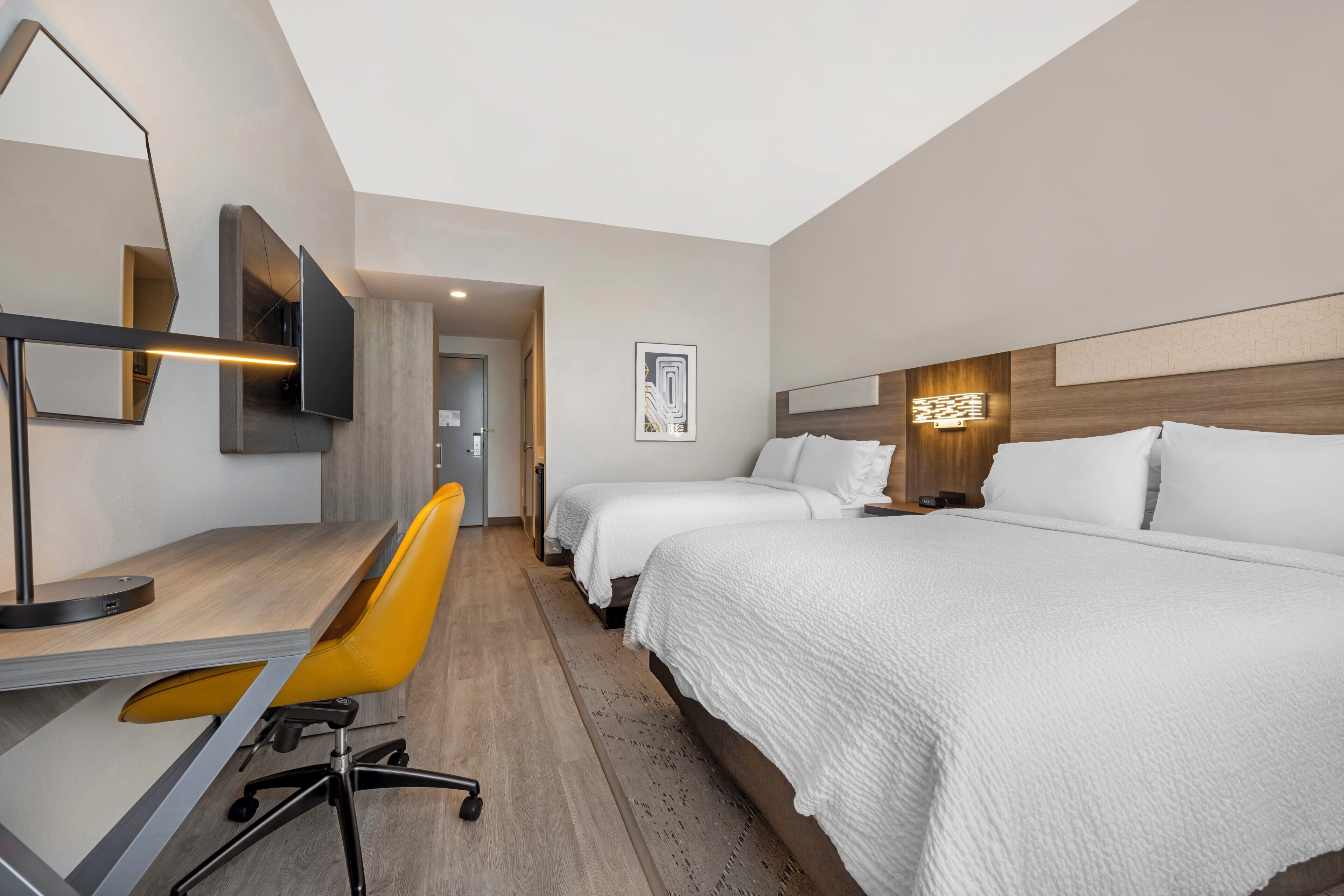 A hotel room with two beds, a desk with a chair, a flat-screen TV, and a modern light fixture.