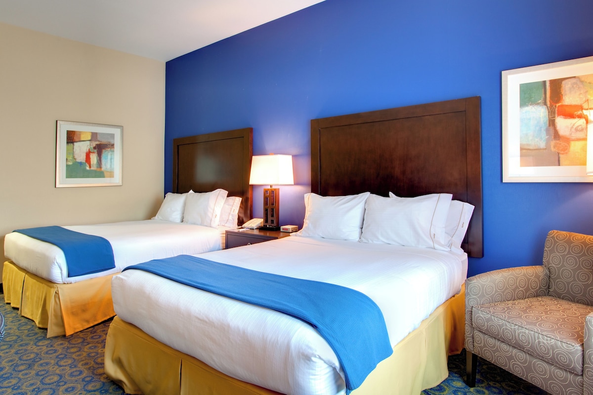 Quarto de hotel com duas camas, mesa de cabeceira com abajur, uma cadeira e obras de arte nas paredes.