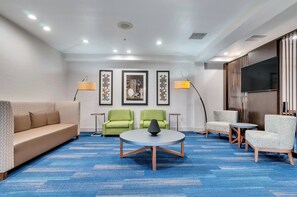 A hotel lobby with a blue carpet, a sofa, two armchairs, a round table, and a TV mounted on the wall. - Holiday Inn Express & Suites, Lake Elsinore by IHG (Lake Elsinore)