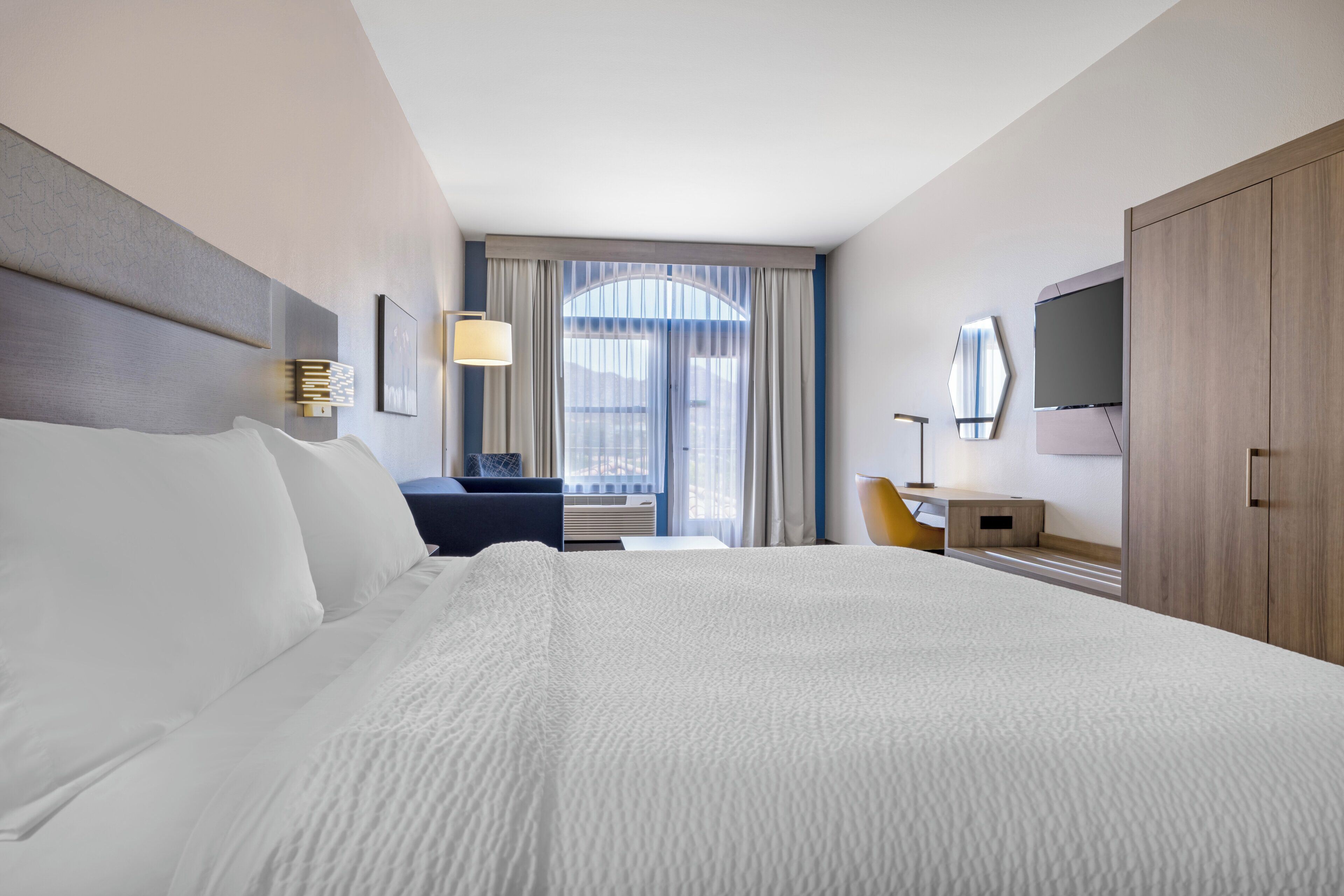 A hotel room with a large bed, a desk, a chair, a TV, and a window with curtains.