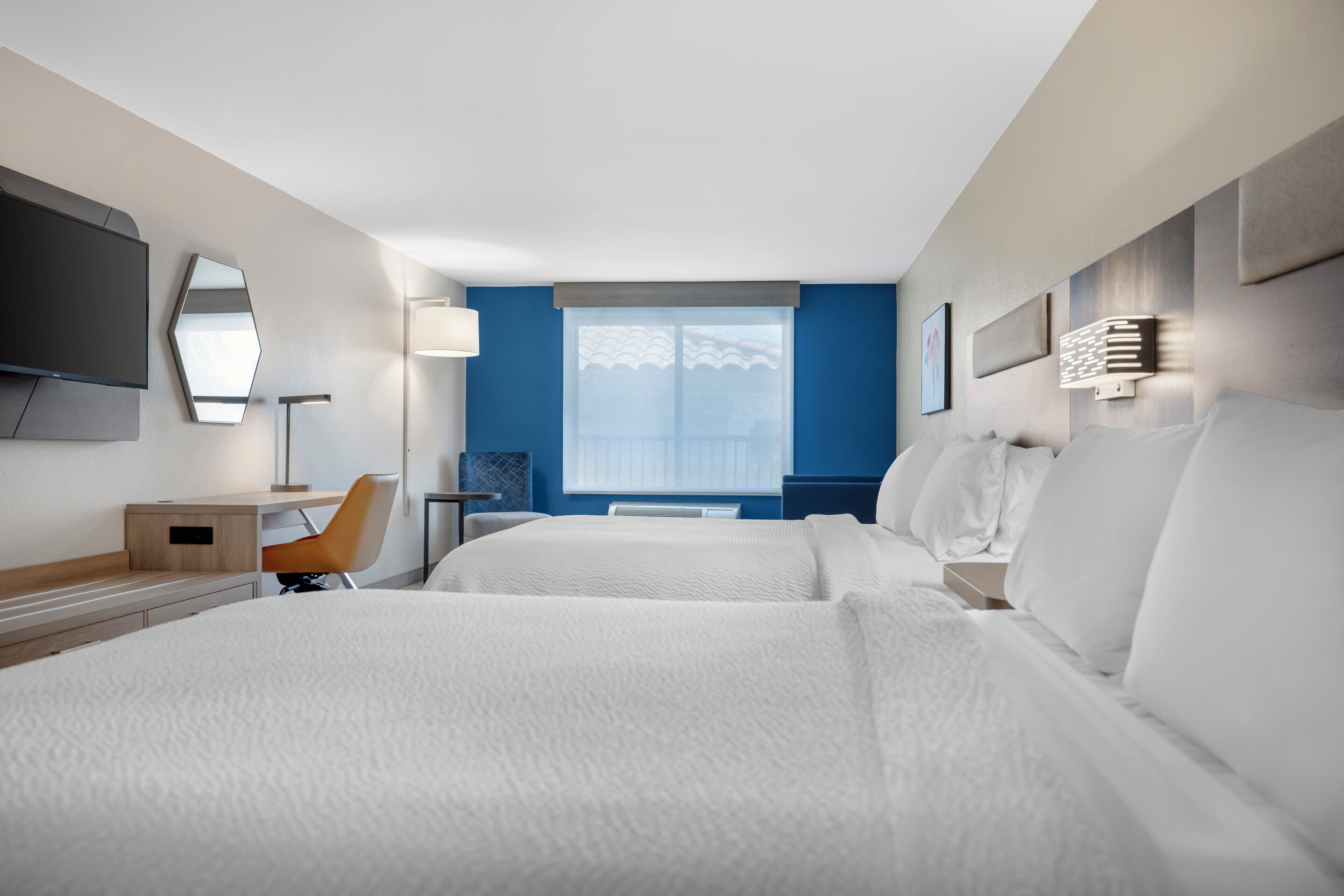 A hotel room with two beds, a desk, a chair, a TV, and a window with blinds.