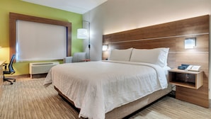 A hotel room with a large bed, a bedside table with a phone and a lamp, a chair, a desk, and a window with blinds.