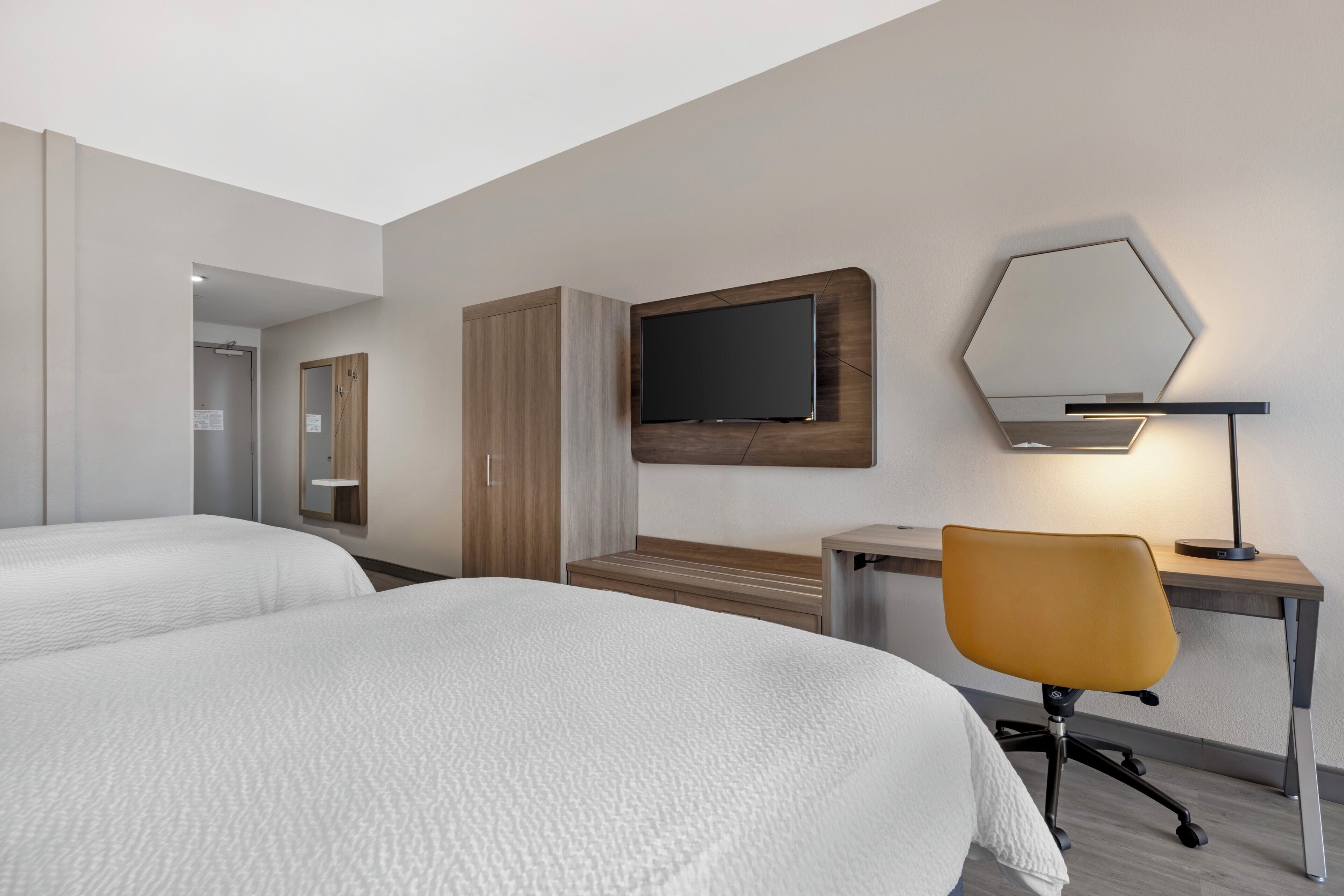 Habitación de hotel con dos camas, un escritorio con silla, un televisor de pantalla plana y un espejo en la pared.