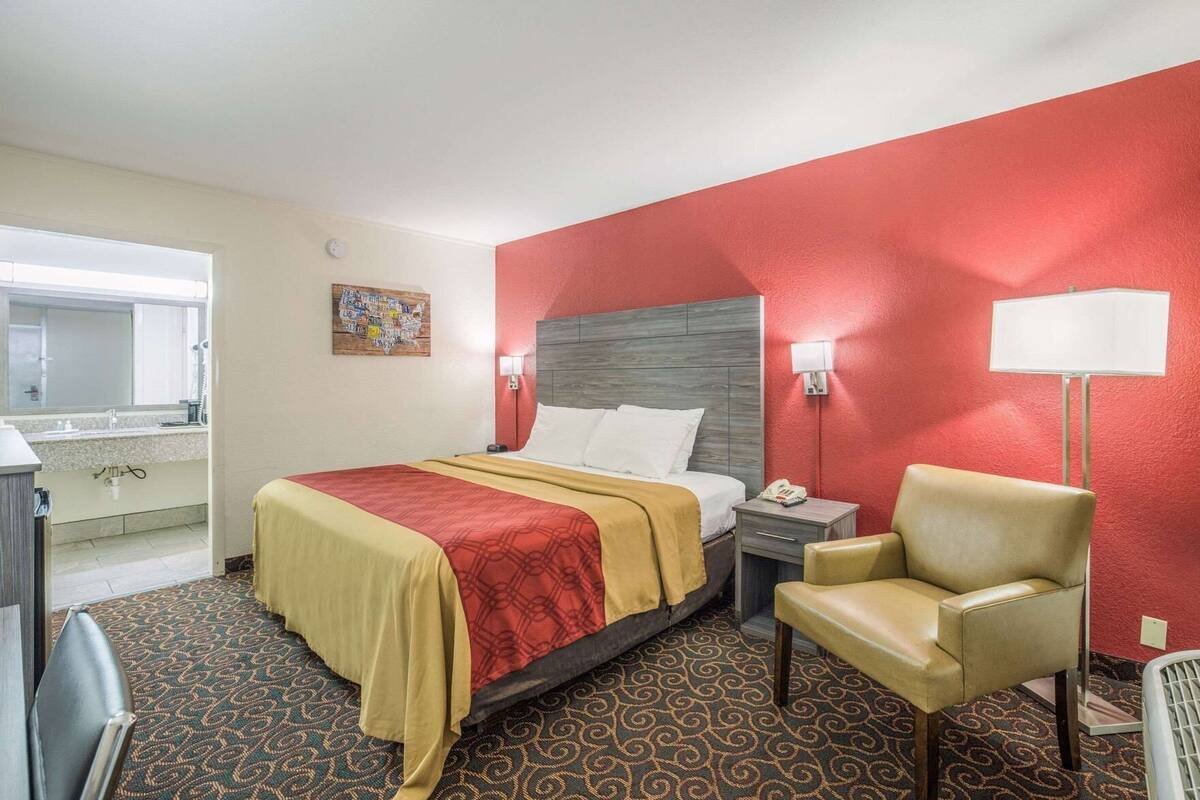 Een hotelkamer met een bed, een stoel, een nachtkastje, een lamp en een badkamer op de achtergrond.
