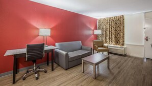 A room with a red wall, a grey sofa, a desk with a chair, a small table, and a window with patterned curtains. - Econo Lodge Thomasville (Thomasville)