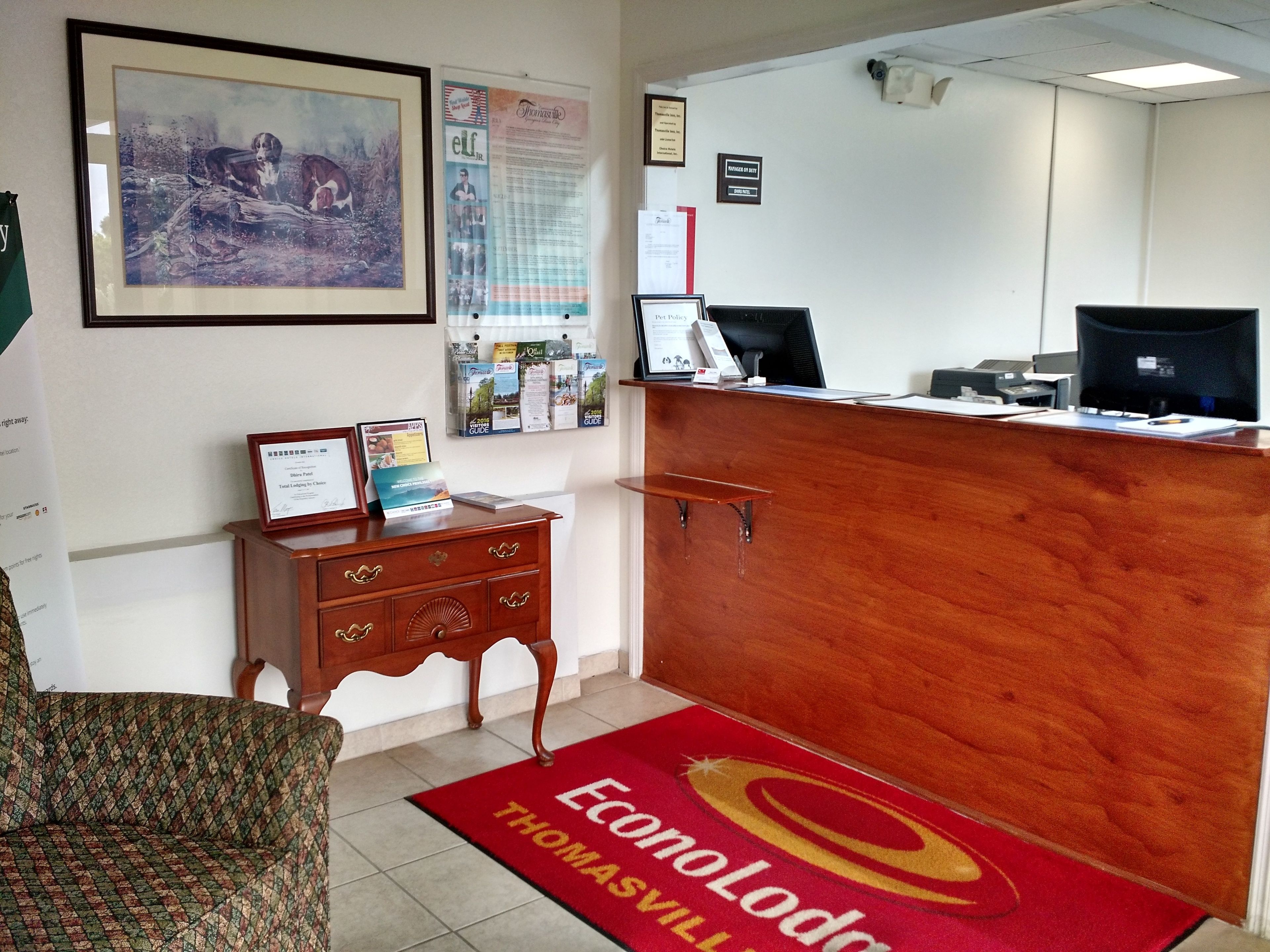A reception area with a wooden desk, a framed certificate, a bulletin board with various notices, and a red carpet with the name 'Thomas' visible.
