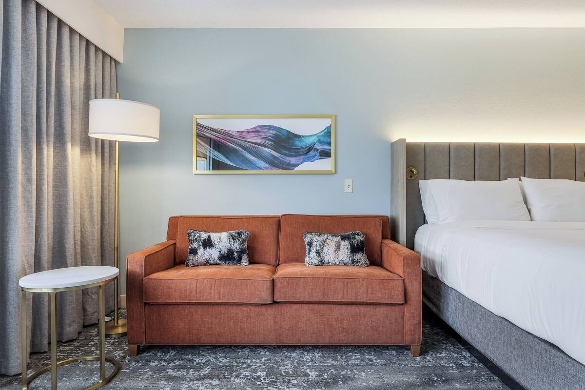 A hotel room with a sofa, a bed, a lamp, and a painting on the wall.