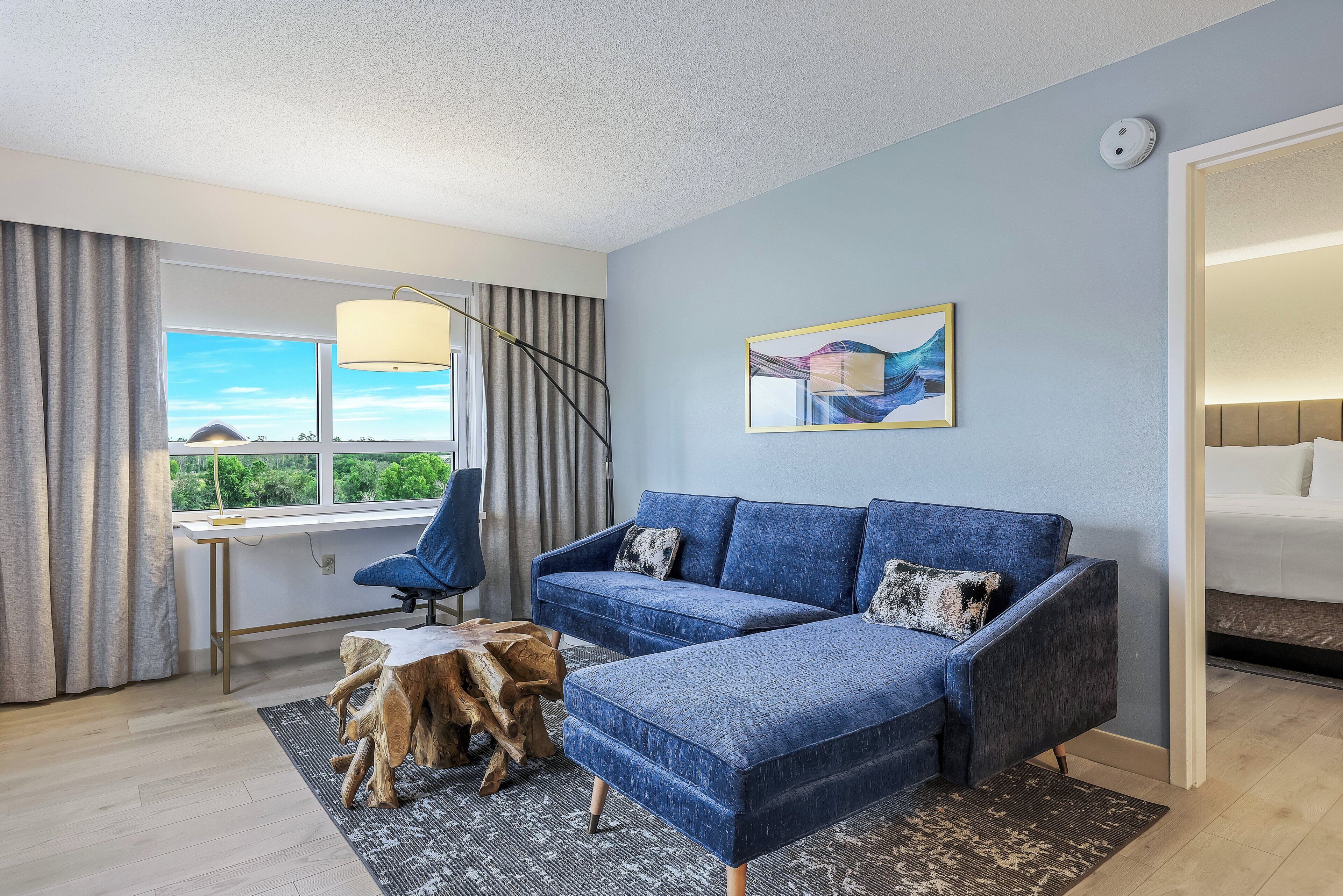 A modern room with a sectional sofa, a desk, a chair, a large window, and a painting on the wall.
