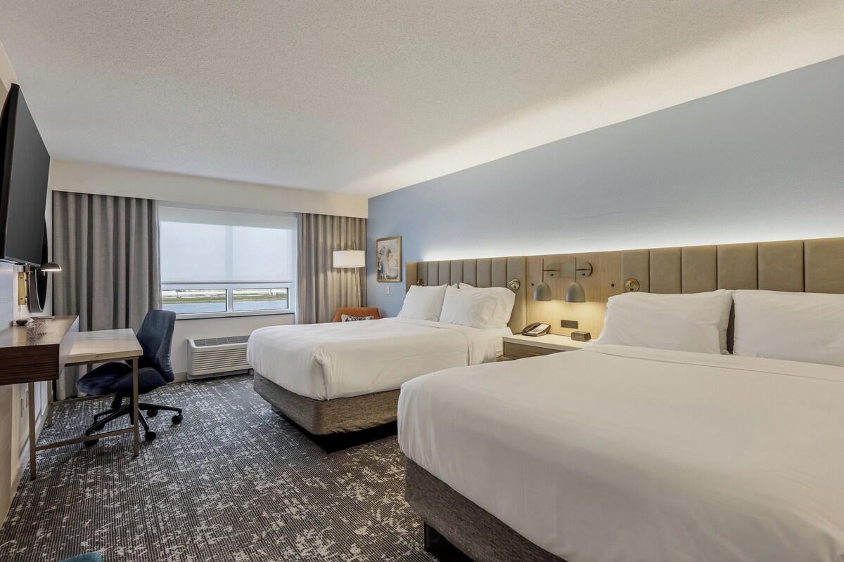 Ein Hotelzimmer mit zwei Betten, einem Schreibtisch, einem Stuhl und einem großen Fenster mit Vorhängen.