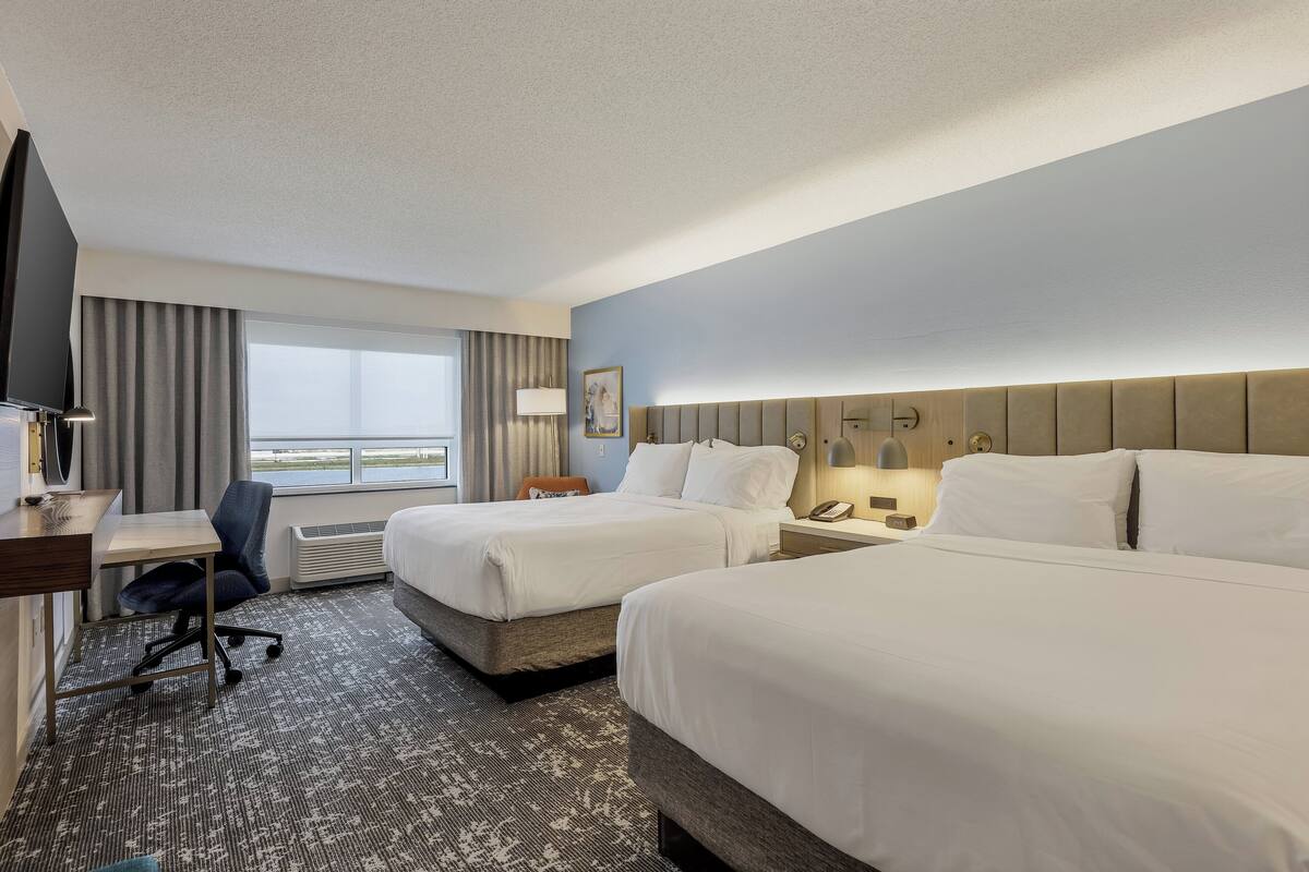 A hotel room with two beds, a desk, a chair, and a large window with curtains.