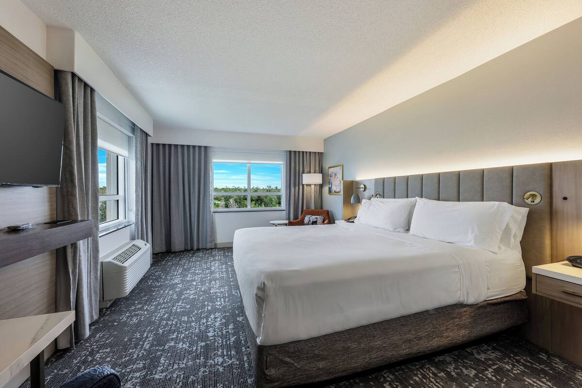 A hotel room with a large bed, a TV, a desk, and a window with a view of the outdoors.