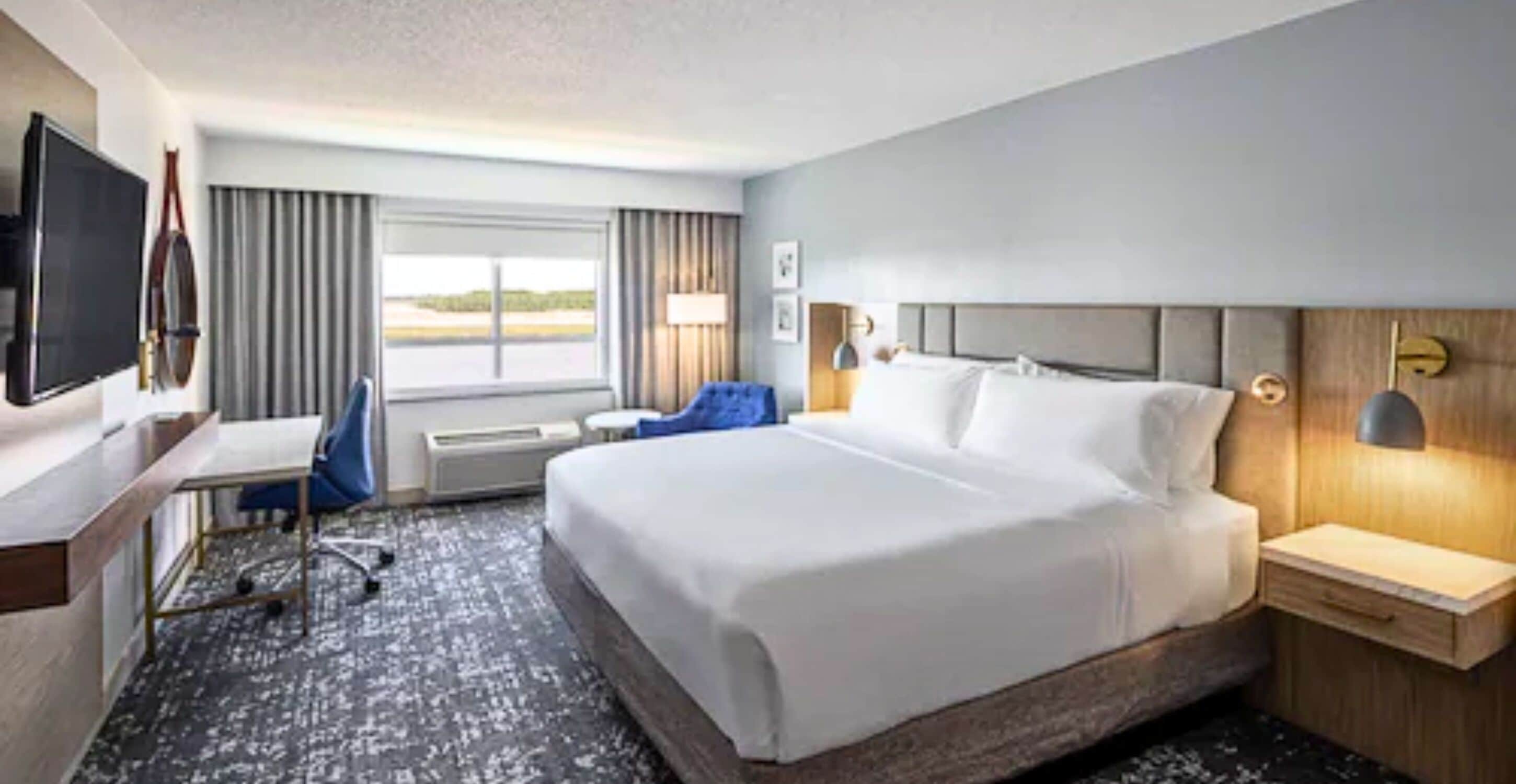 A hotel room with a large bed, a desk with a chair, a TV, and a window with curtains.