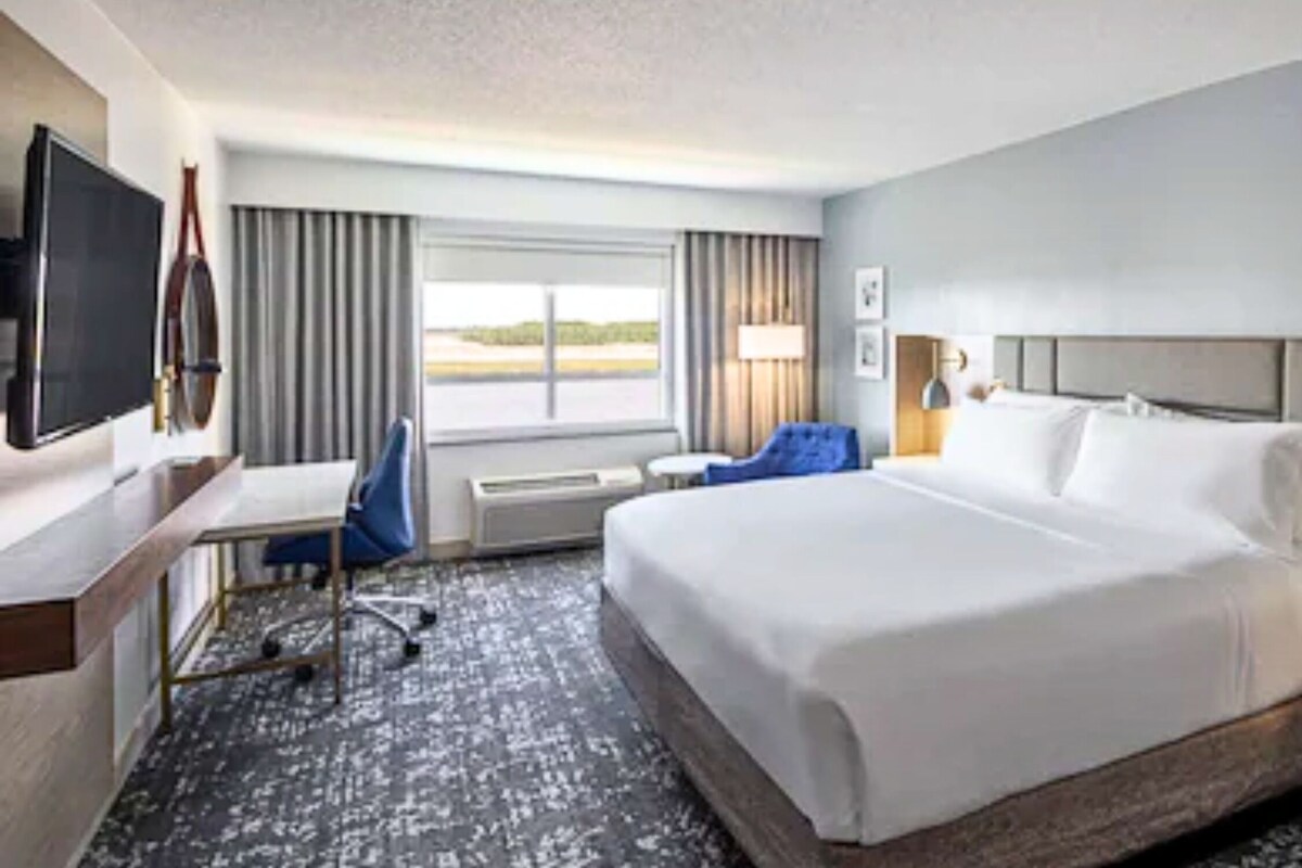 Habitación de hotel con una cama grande, un escritorio con silla, un televisor y una ventana con cortinas.