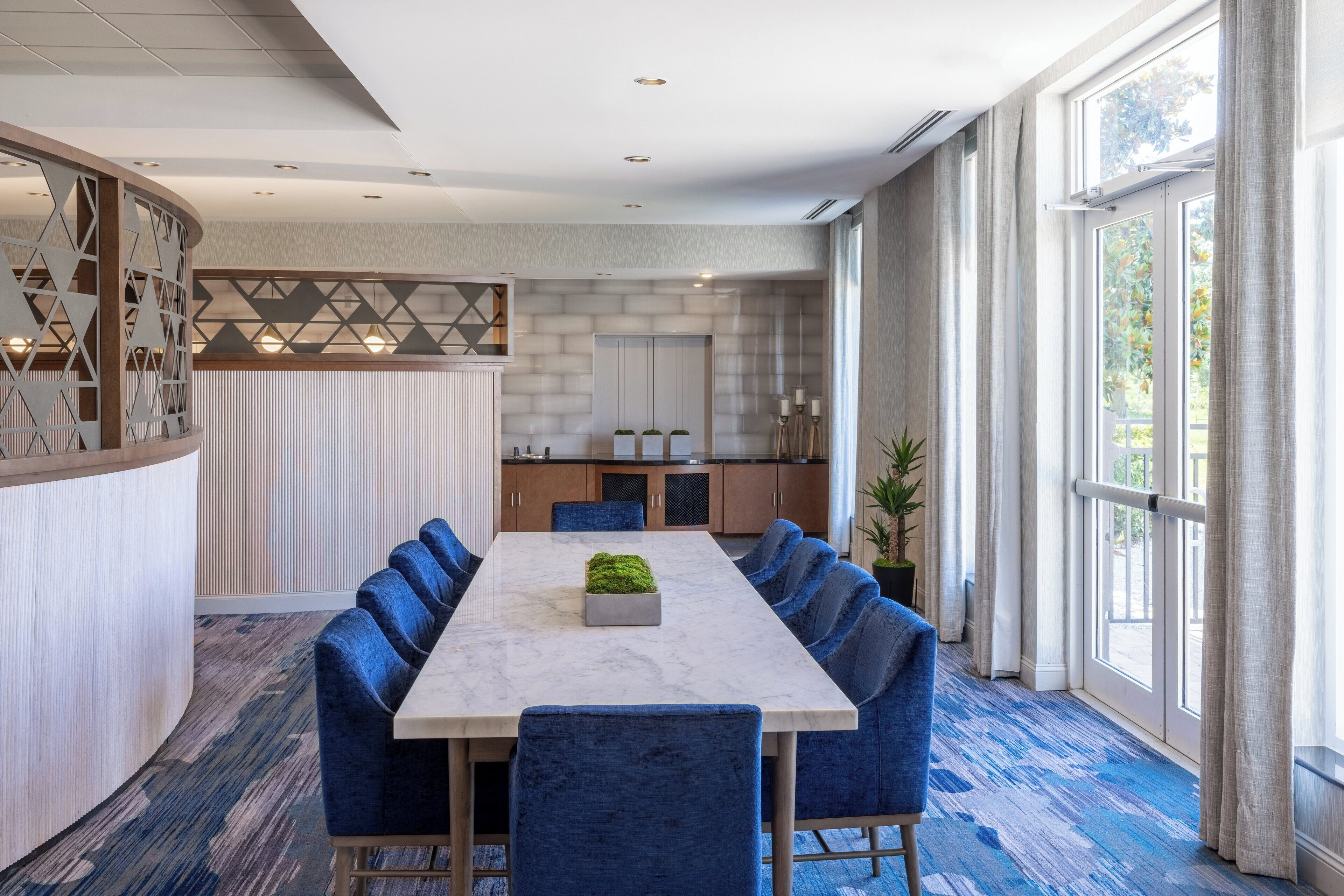 A modern dining area with a long table, blue upholstered chairs, a marble table, and a kitchen in the background.