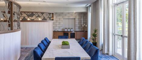 A modern dining area with a long table, blue upholstered chairs, a marble table, and a kitchen in the background.