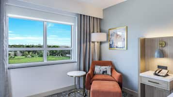 Habitación de hotel con un ventanal grande, una mesita redonda pequeña, un sillón naranja, una mesita de noche con lámpara y un cuadro en la pared.