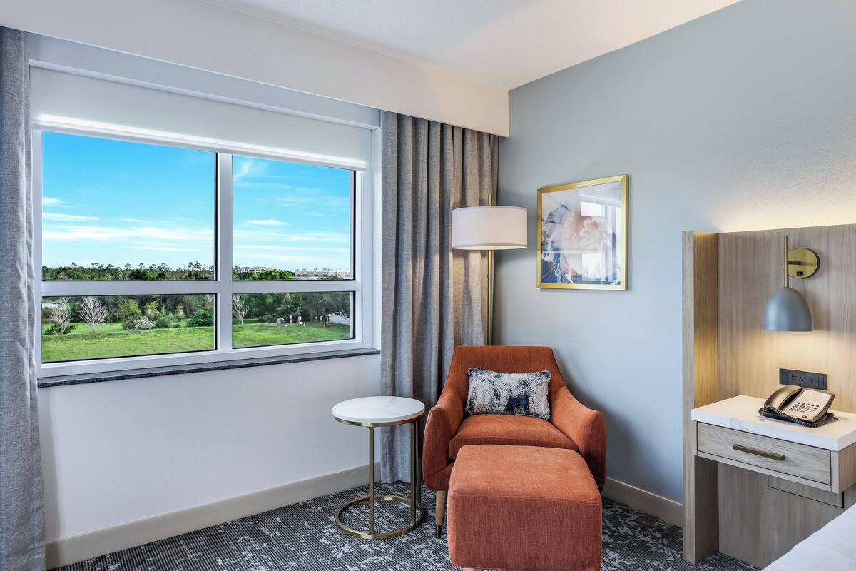 A hotel room with a large window, a small round table, a sofa, a bedside table with a lamp, and a painting on the wall.