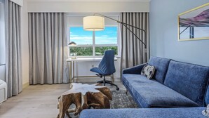 A modern living room with a blue sofa, a wooden coffee table, a desk with a chair, and a large window with curtains.