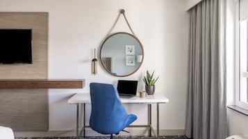 Quarto de hotel com uma escrivaninha branca, um laptop, uma cadeira azul, um espelho redondo, uma TV de tela plana, uma planta em vaso e uma janela com cortinas.