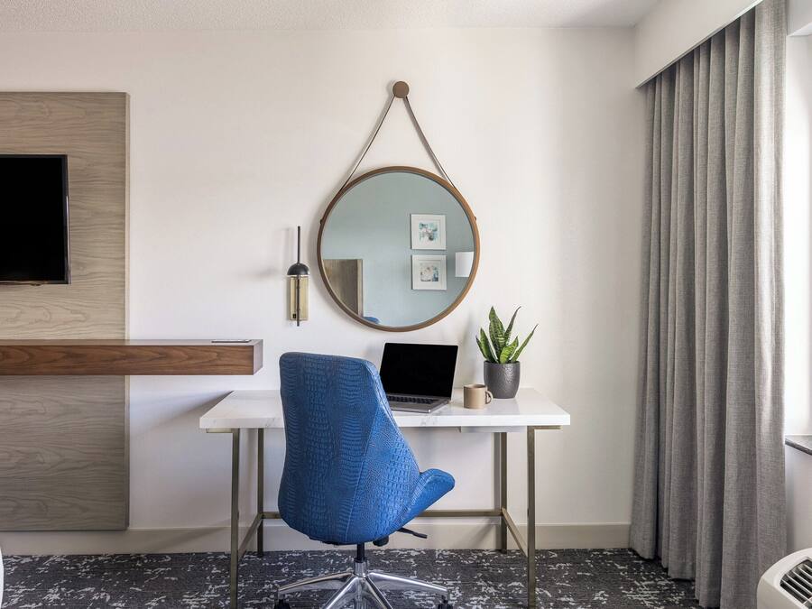 Quarto de hotel com uma escrivaninha branca, um laptop, uma cadeira azul, um espelho redondo, uma TV de tela plana, uma planta em vaso e uma janela com cortinas.