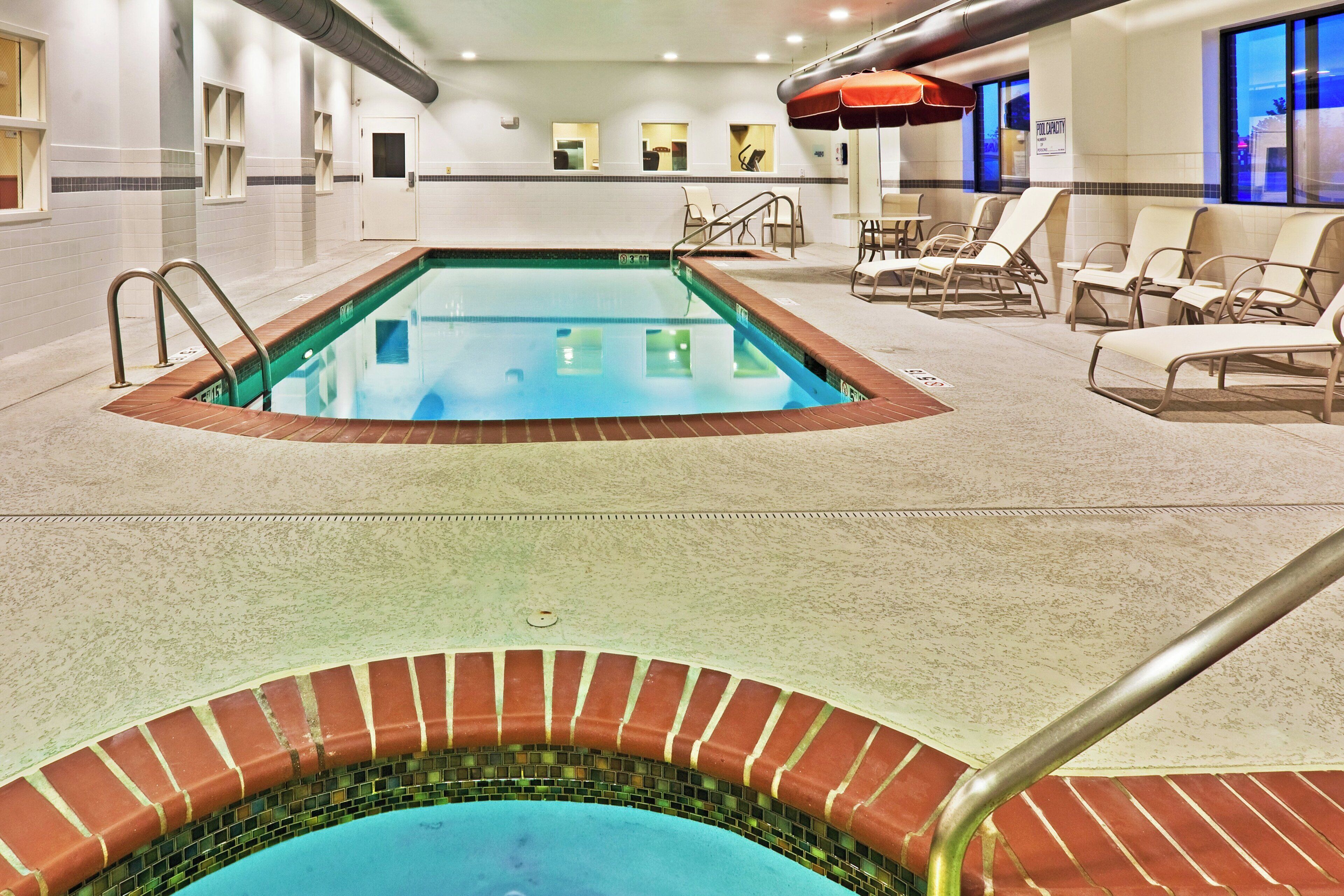 An indoor swimming pool with a tiled edge, lounge chairs, and a pool capacity sign.