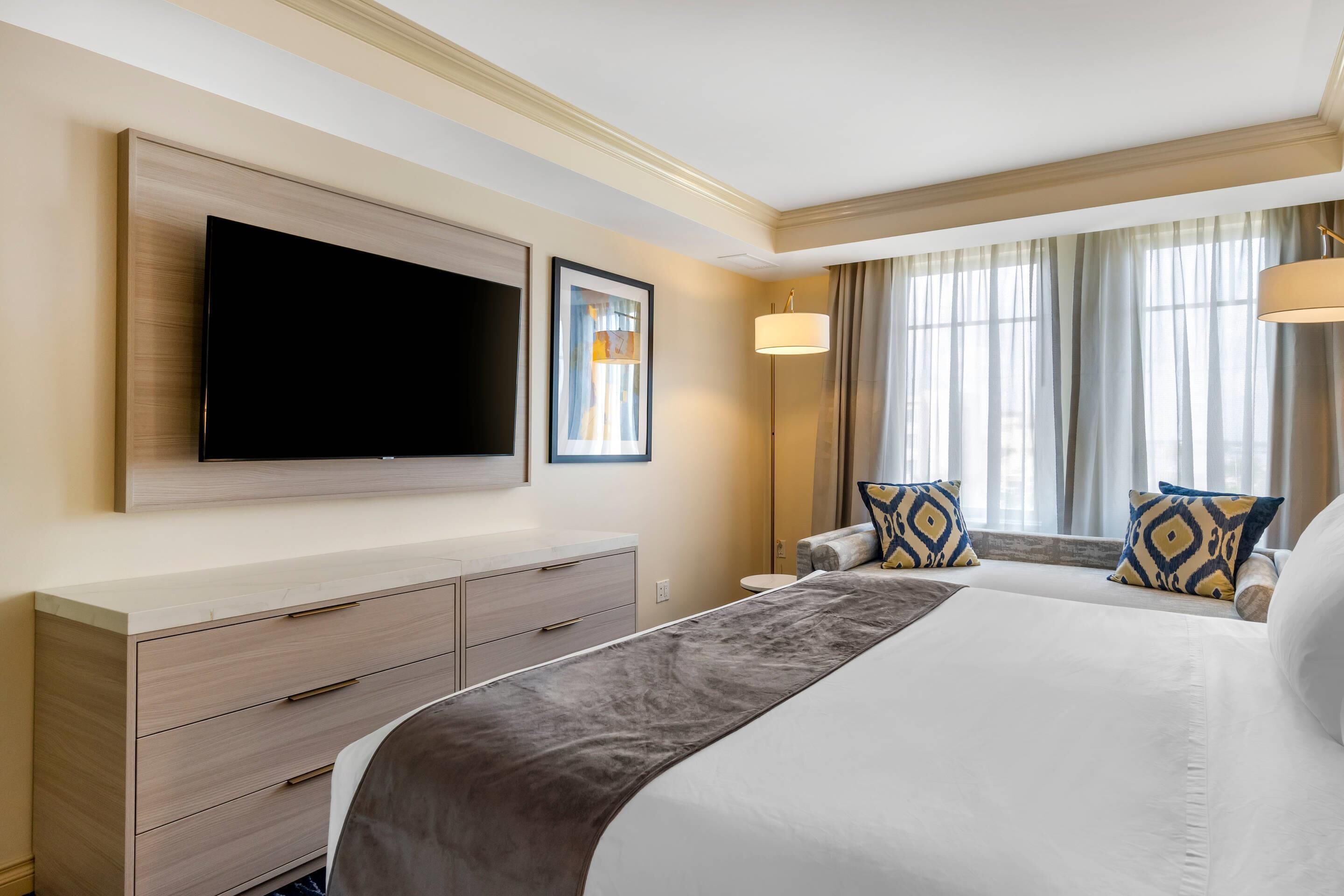 A hotel room with a large bed, a flat-screen TV mounted on the wall, a bedside table, and a window with curtains.