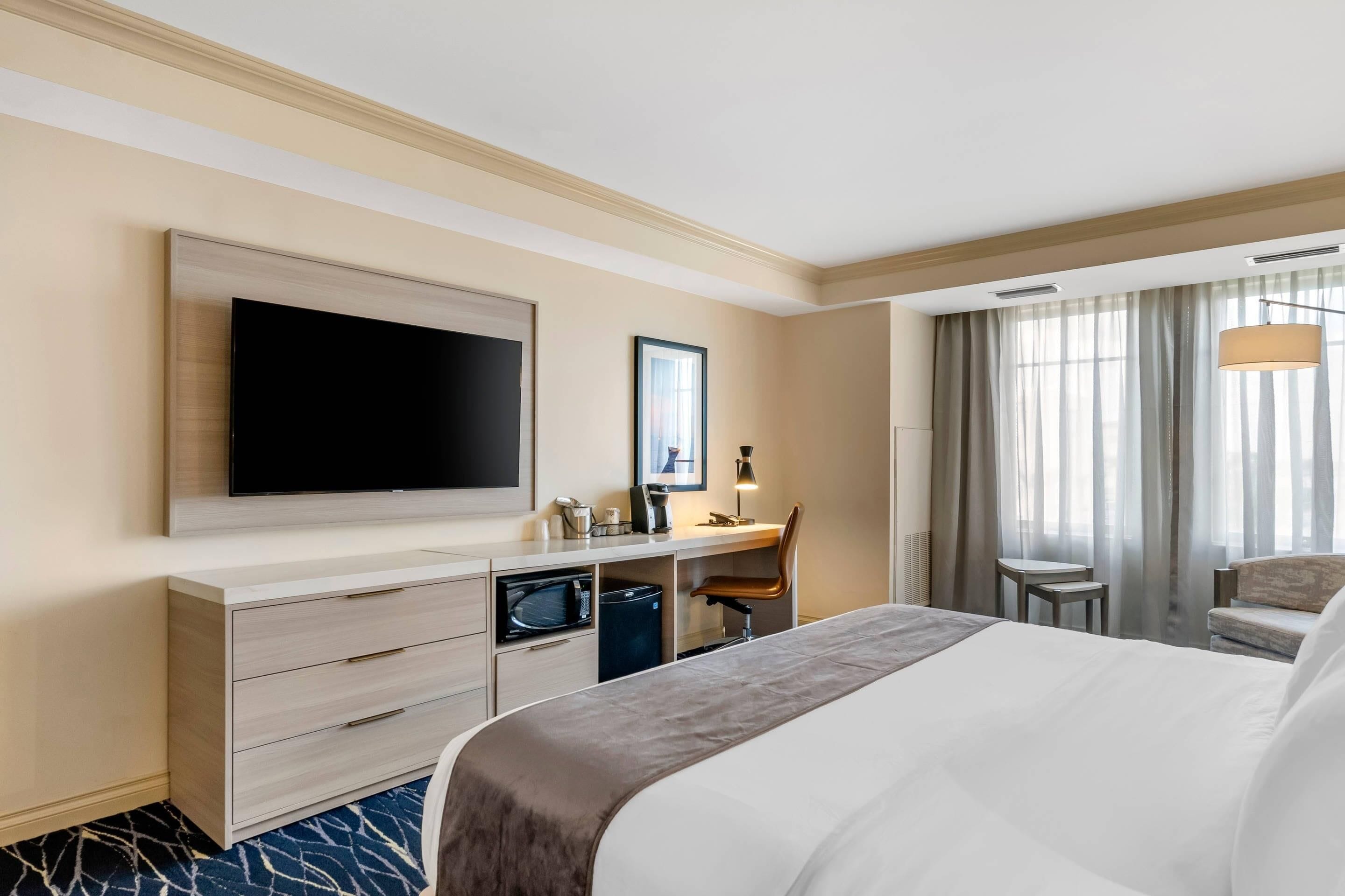 A hotel room with a large bed, a desk with a computer, a flat-screen TV, a chair, and a window with curtains.