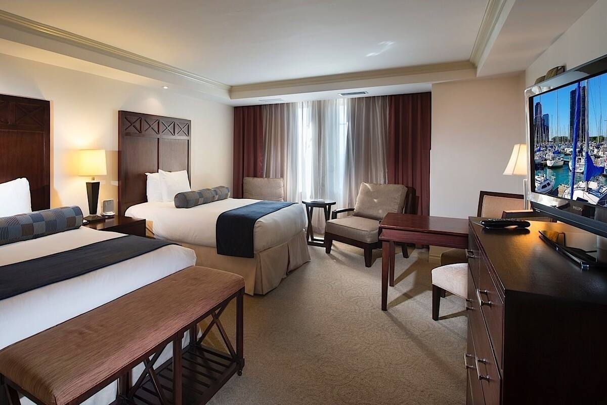 Habitación de hotel con dos camas, un escritorio, una silla, un televisor y vista al puerto deportivo.
