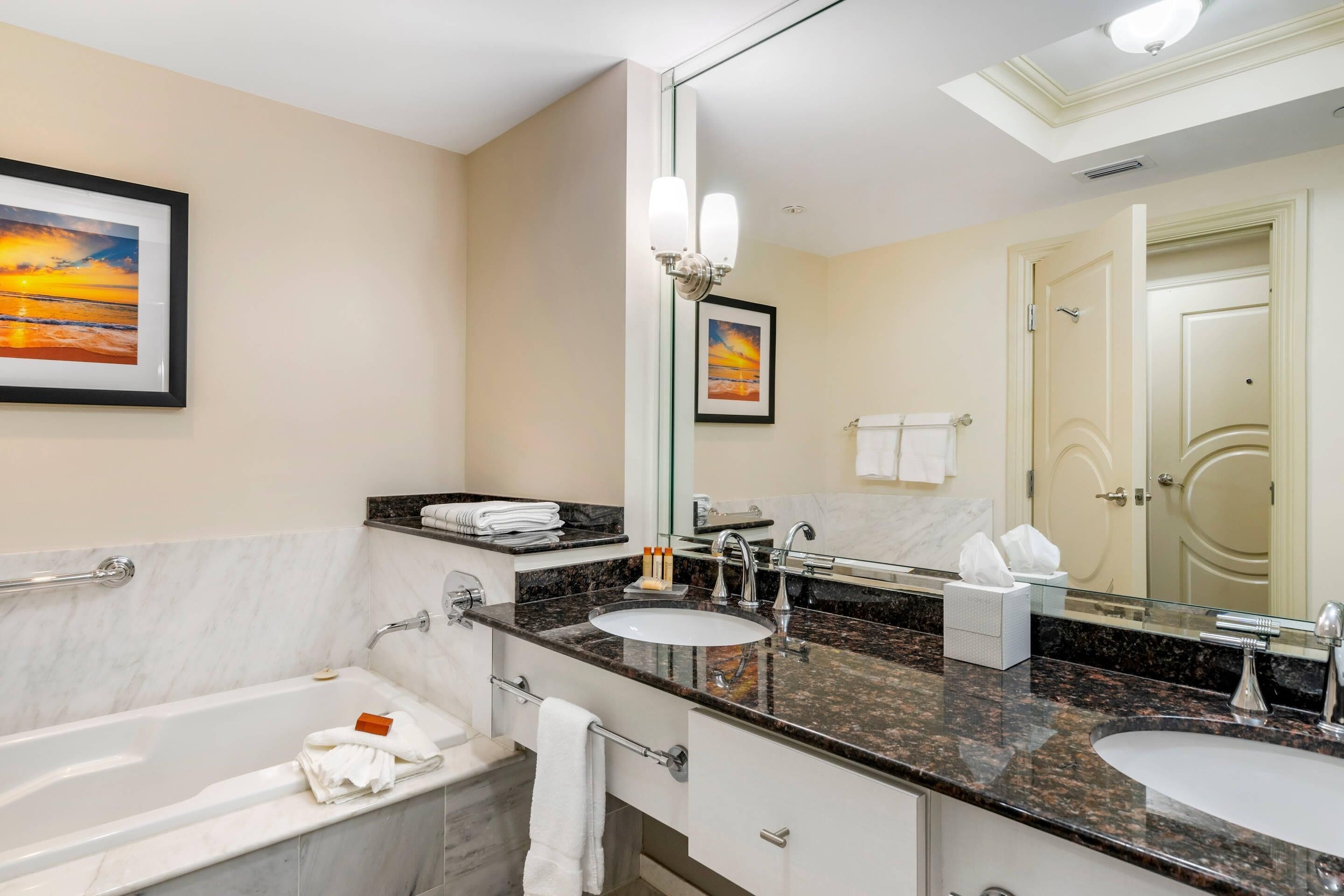 A modern bathroom with a marble sink, a bathtub, and a large mirror. There are towels and toiletries on the counter.