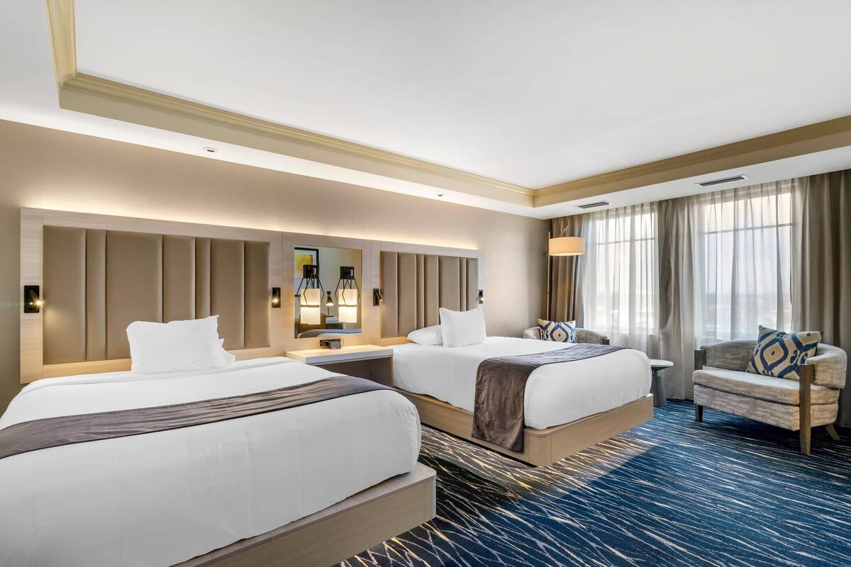Habitación de hotel con dos camas, un sofá, un espejo grande y vistas al exterior.