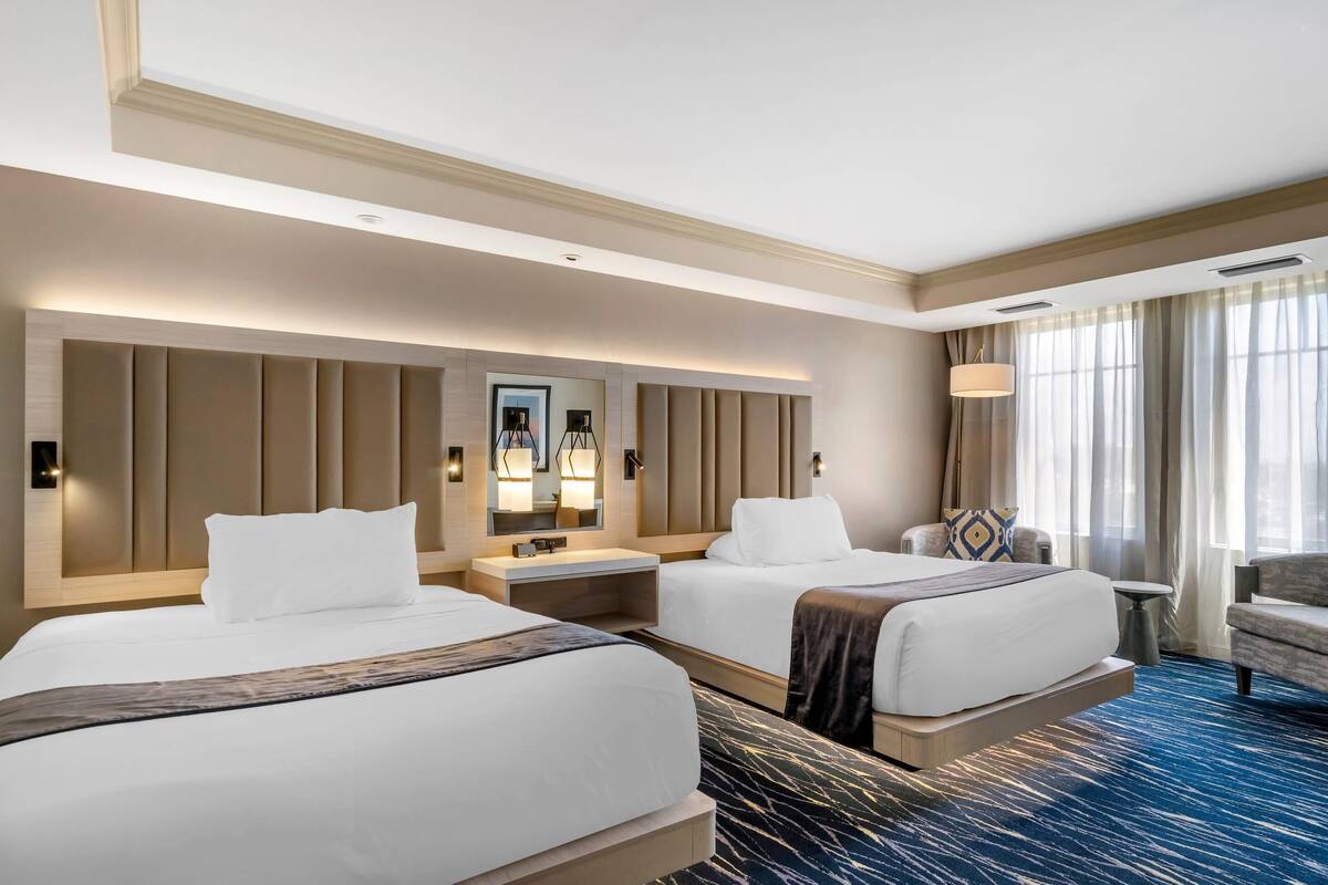 Habitación de hotel con dos camas, un espejo grande, una mesita de noche, una silla y vista al exterior.
