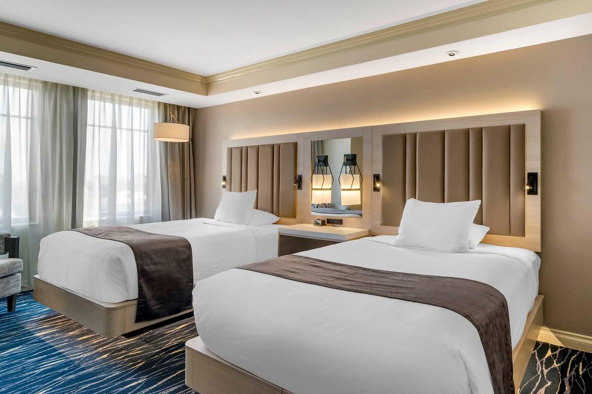 Habitación de hotel con dos camas, un escritorio, una silla y un amplio ventanal con cortinas.