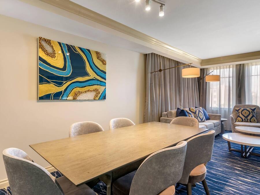 A hotel room with a dining area featuring a wooden table and chairs, a sofa, and a large abstract painting on the wall.