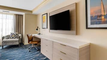 A hotel room with a large flat-screen TV mounted on the wall, a desk with drawers, a comfortable armchair, and a large window with curtains.