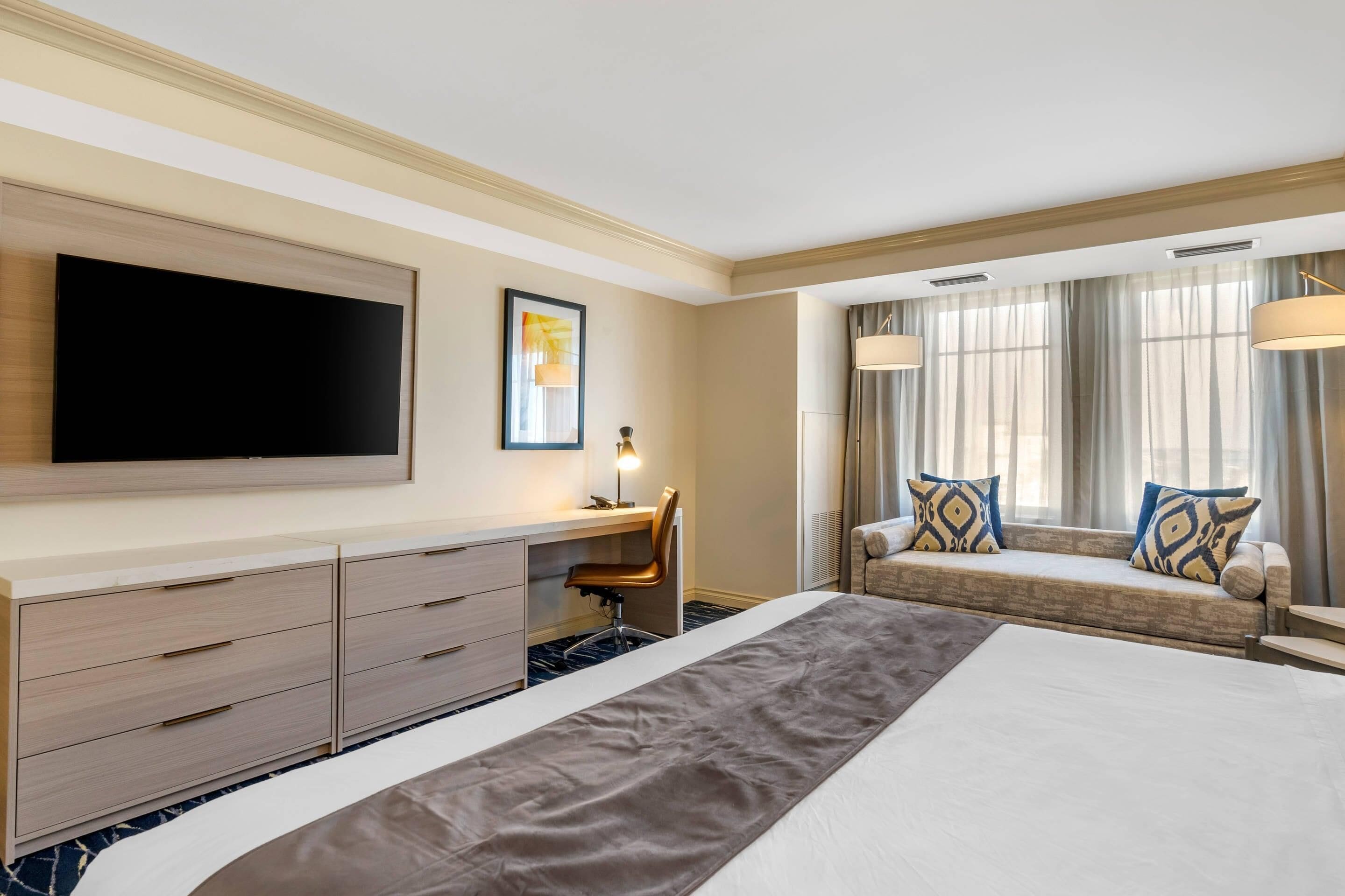 Habitación de hotel con una cama grande, un escritorio con silla, un televisor de pantalla plana, una ventana con cortinas y un sofá con cojines.