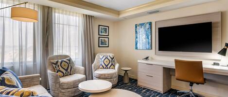 A hotel room with a large flat-screen TV, two armchairs, a sofa, a round coffee table, a desk with a chair, and a patterned carpet.