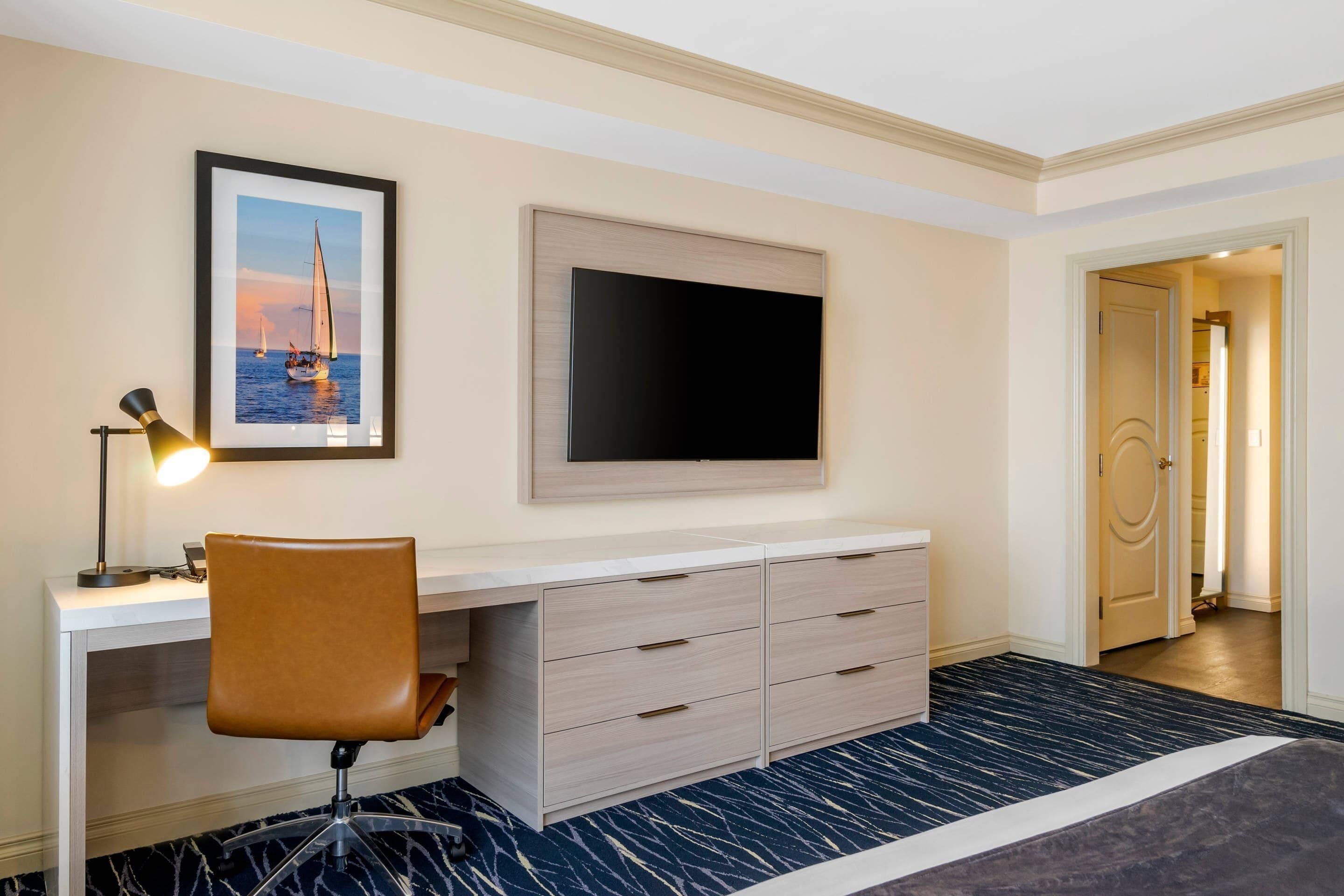 A hotel room with a desk, chair, TV, and a framed picture of a sailboat.