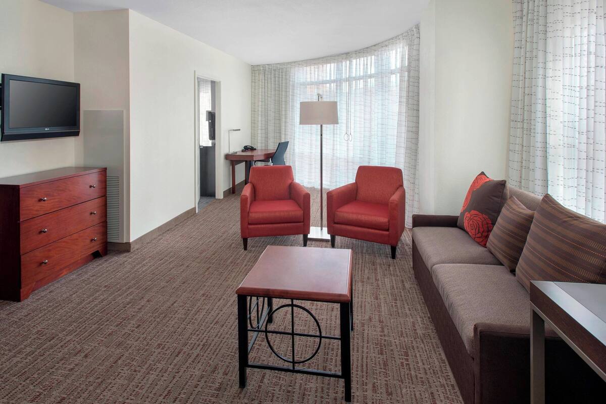 Habitación de hotel con sofá, dos sillones, una mesita, televisor montado en la pared, escritorio con silla y lámpara.