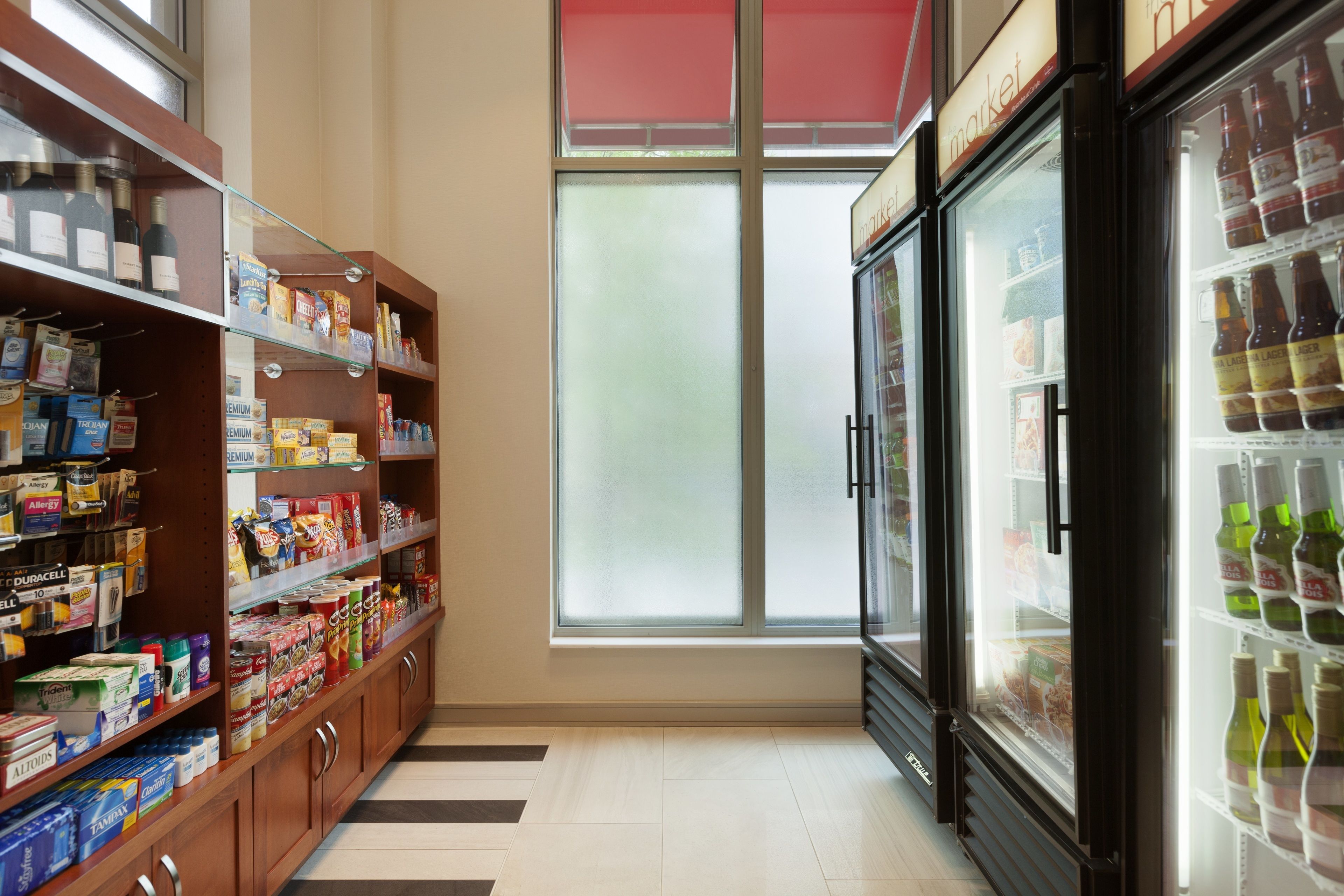 Un pasillo de supermercado con estantes llenos de diversos productos, una sección refrigerada con bebidas, y un ventanal que proporciona luz natural.