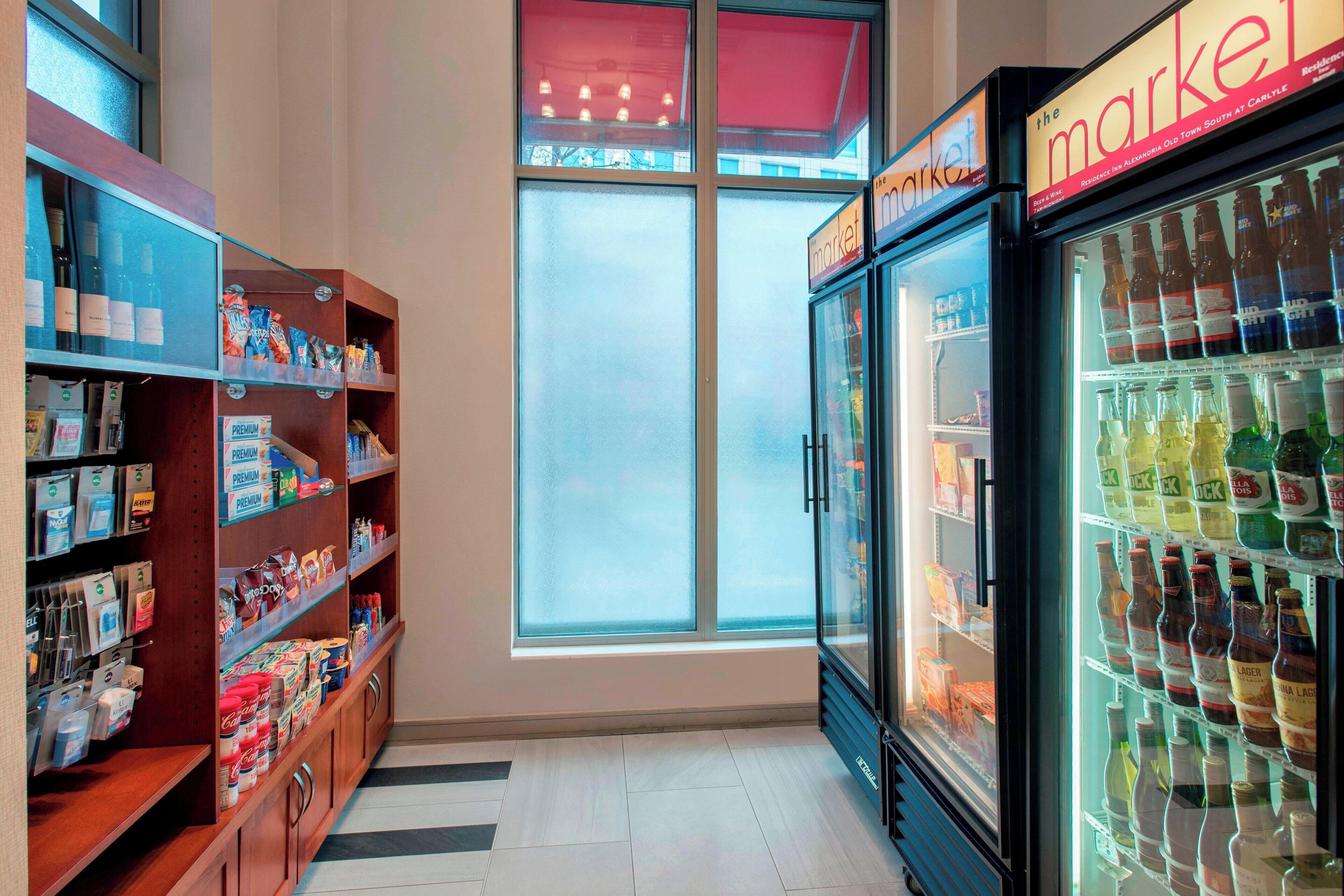 Interior de una tienda con estantes llenos de productos diversos, una sección refrigerada con bebidas y un amplio ventanal que permite la entrada de luz natural.