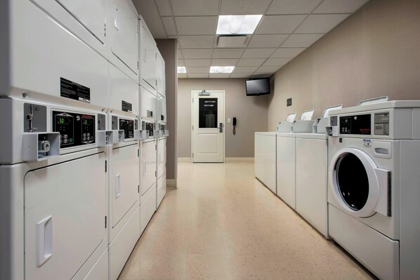 A laundry facility with multiple washing machines and dryers, a flat-screen TV mounted on the wall, and a door leading to another room.